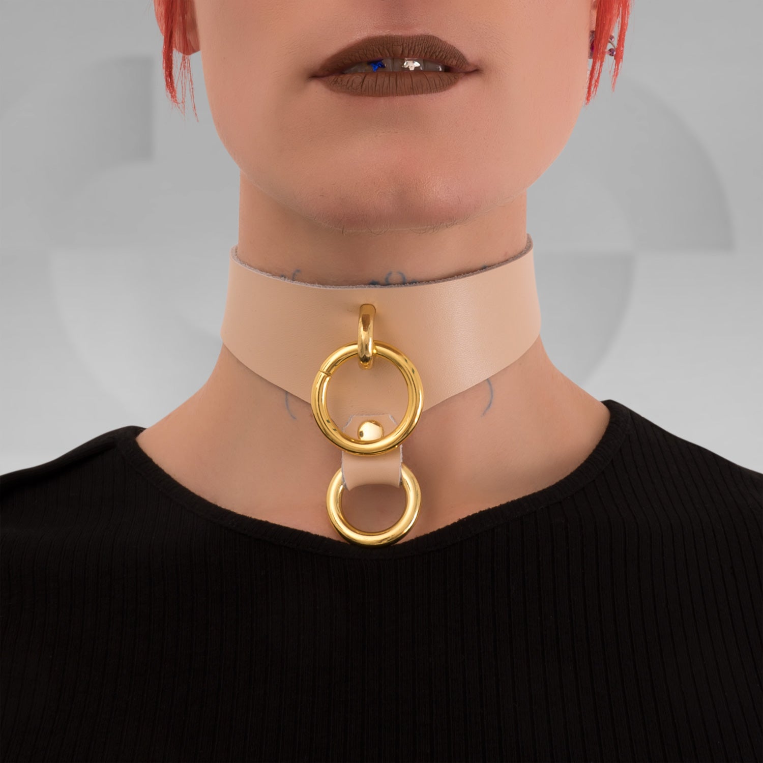This image features a model wearing a chic nude leather choker that highlights its sophisticated design. The choker is adorned with prominent gold rings, creating a striking focal point that beautifully accentuates the neckline. Its elegant hue complements various styles, making it a versatile accessory for both casual outings and bold nightlife looks.