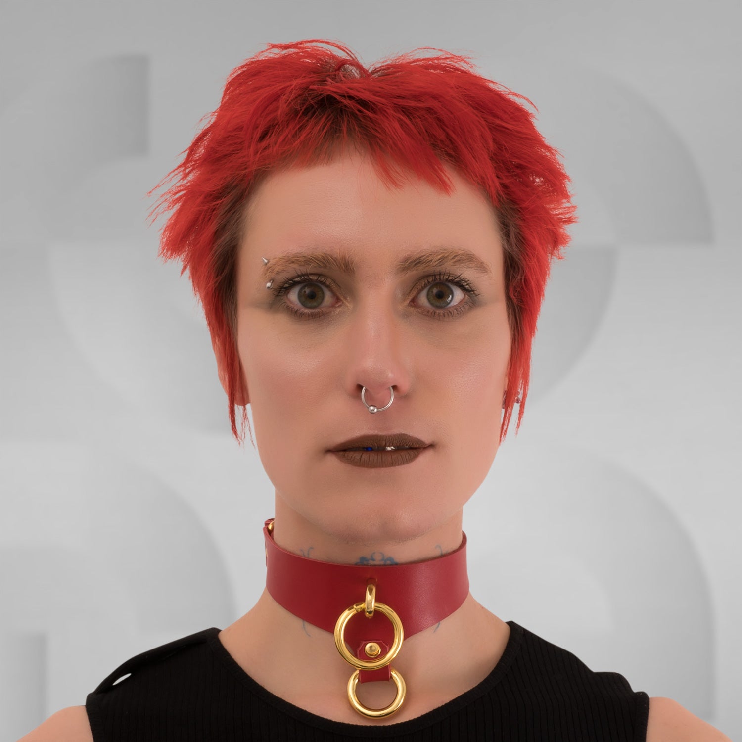 This image features a model wearing a striking red leather choker that showcases its bold design and contemporary flair. The choker features a prominent gold ring at the front, creating an eye-catching focal point that enhances the model's neckline and adds a touch of luxury to her look.