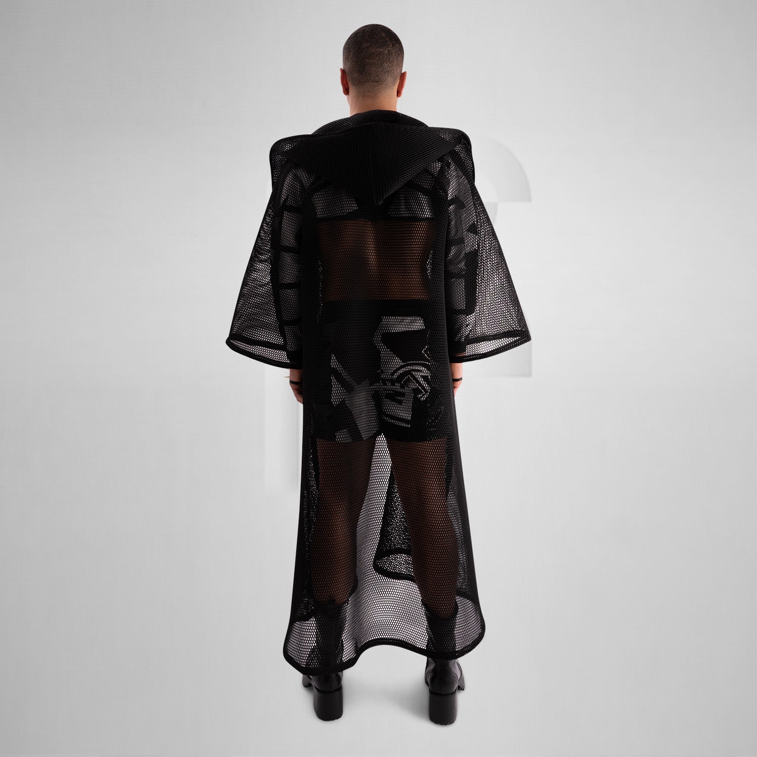 A model wearing a sheer black top with a dramatic black mesh floor-length robe, accessorized with a metallic collar, epitomizing avant-garde fashion and clubwear style, against a neutral gray background.
