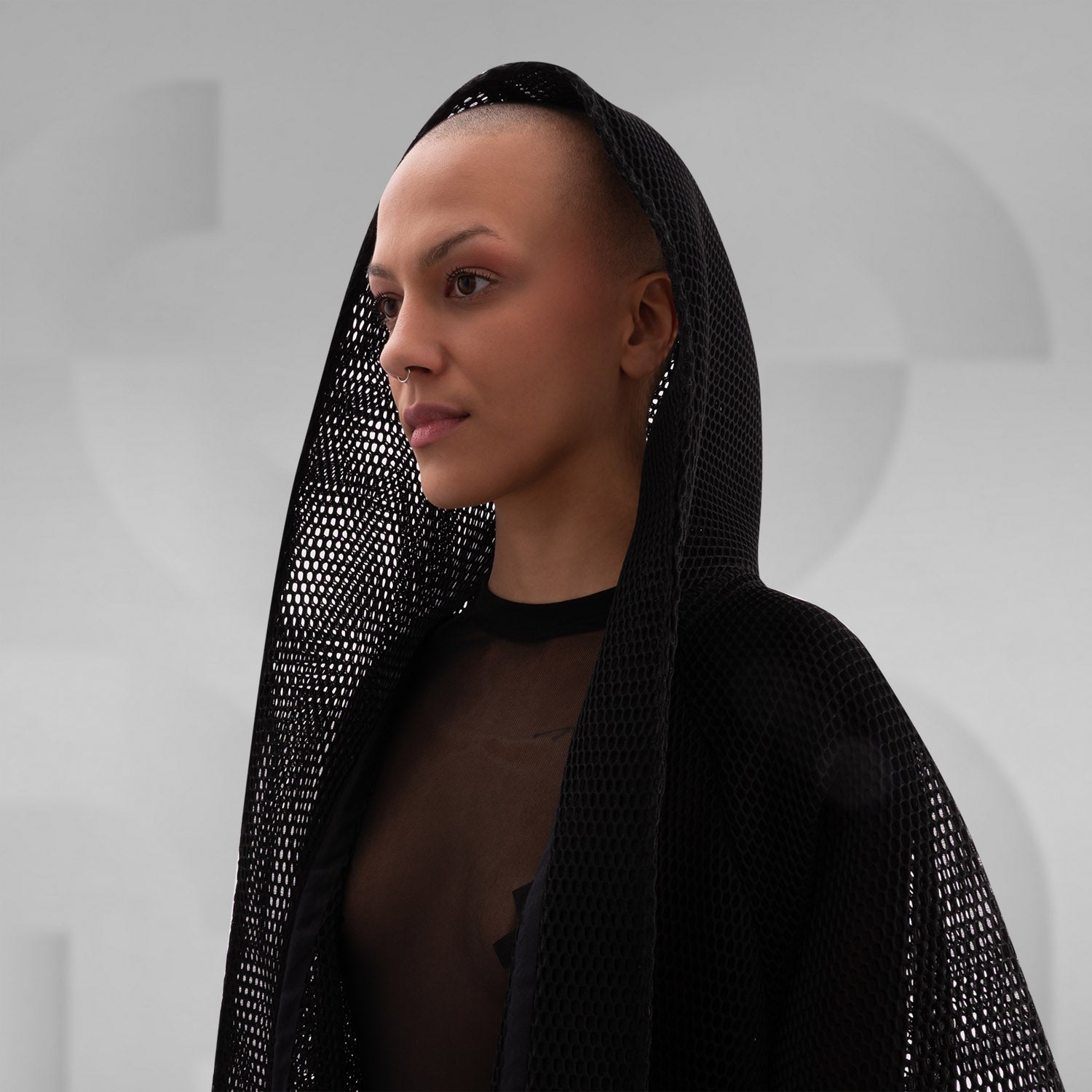 A model wearing a sheer black top with a dramatic black mesh floor-length robe, accessorized with a metallic collar, epitomizing avant-garde fashion and clubwear style, against a neutral gray background.