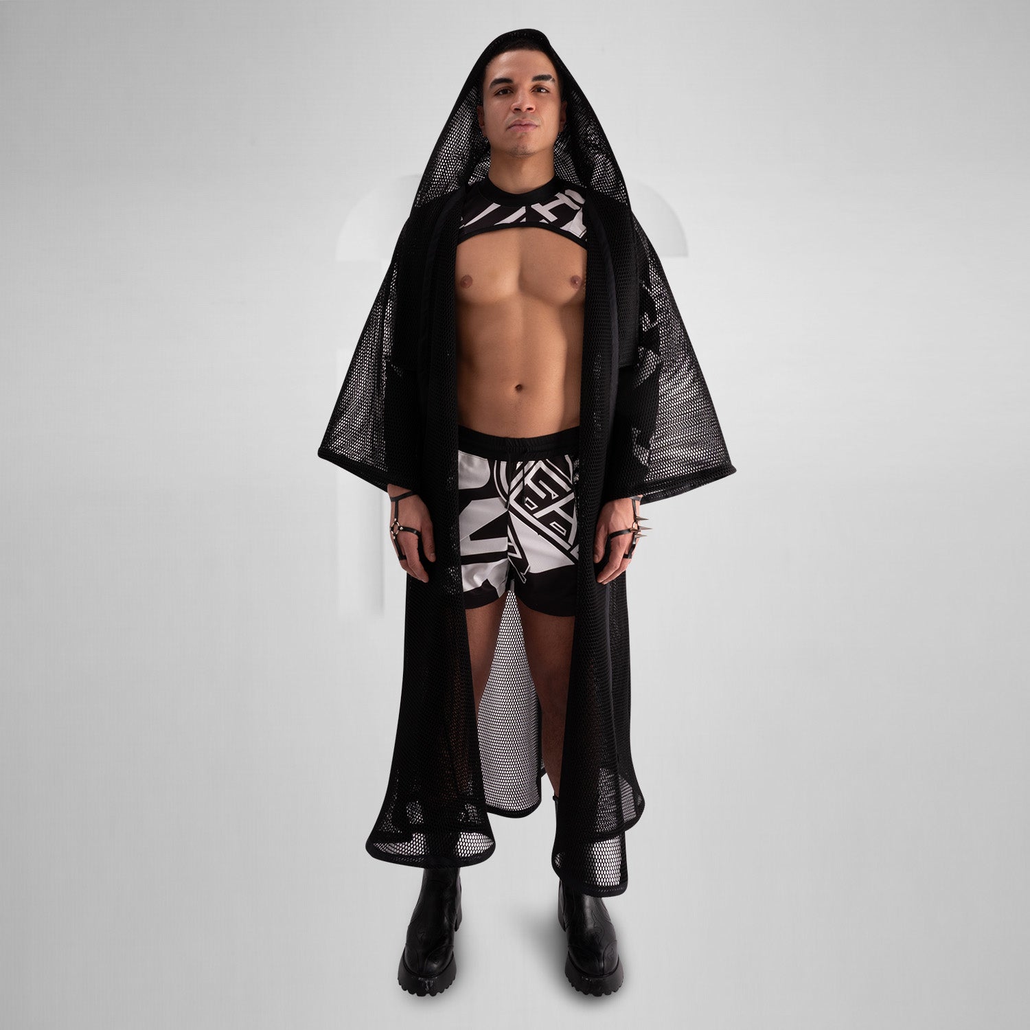 A model wearing a sheer black top with a dramatic black mesh floor-length robe, accessorized with a metallic collar, epitomizing avant-garde fashion and clubwear style, against a neutral gray background.