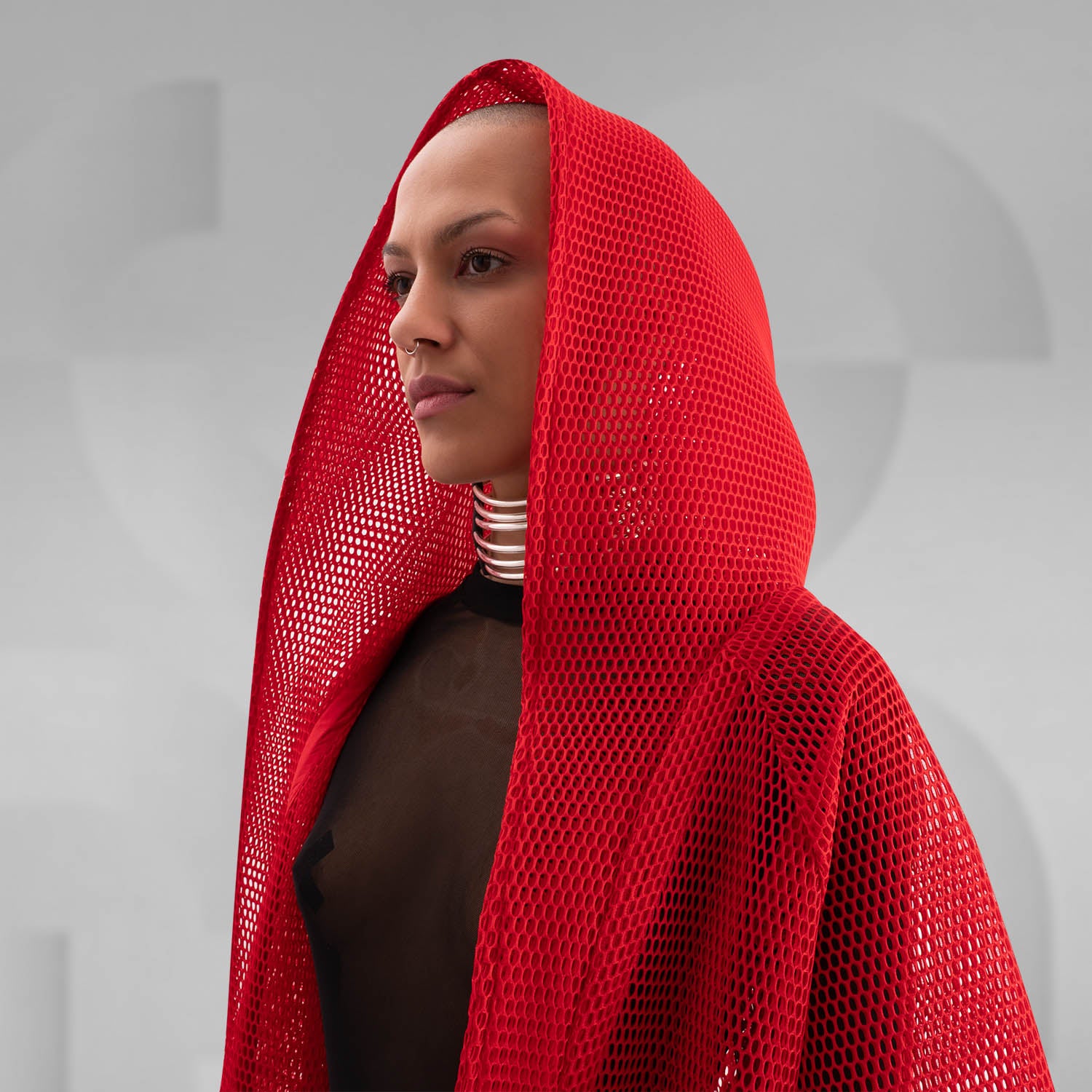 A model wearing a sheer black top with a dramatic red mesh floor-length robe, accessorized with a metallic collar, epitomizing avant-garde fashion and clubwear style, against a neutral gray background.