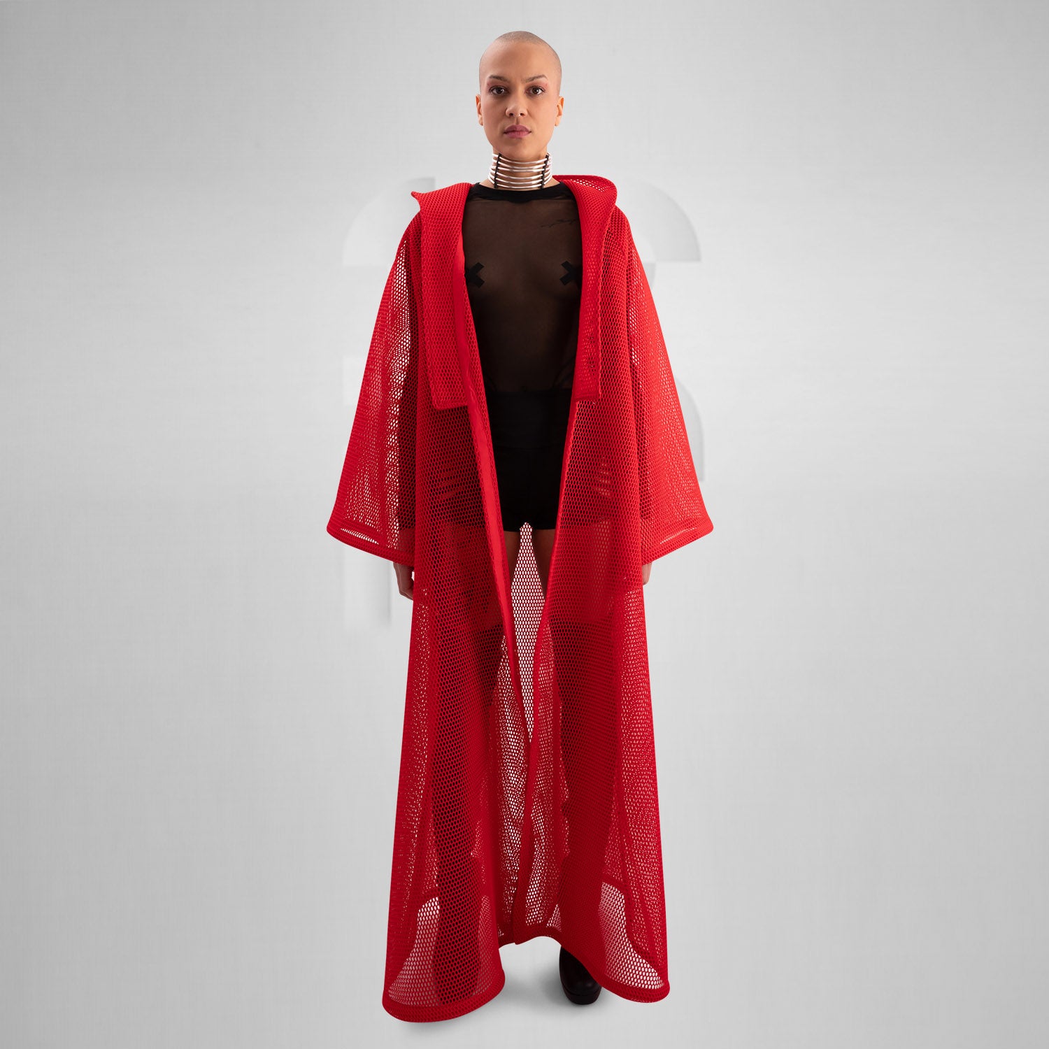 A model wearing a sheer black top with a dramatic red mesh floor-length robe, accessorized with a metallic collar, epitomizing avant-garde fashion and clubwear style, against a neutral gray background.