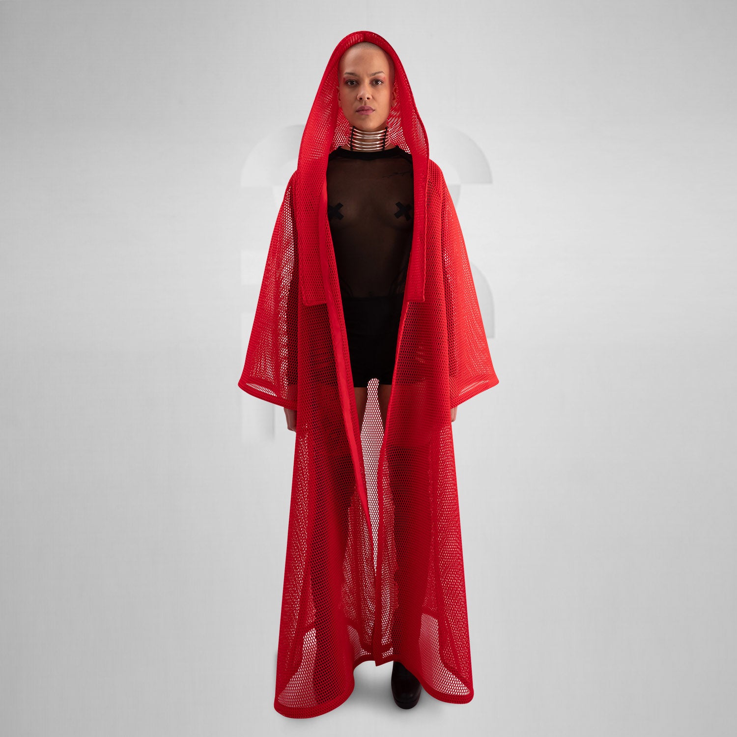 A model wearing a sheer black top with a dramatic red mesh floor-length robe, accessorized with a metallic collar, epitomizing avant-garde fashion and clubwear style, against a neutral gray background.