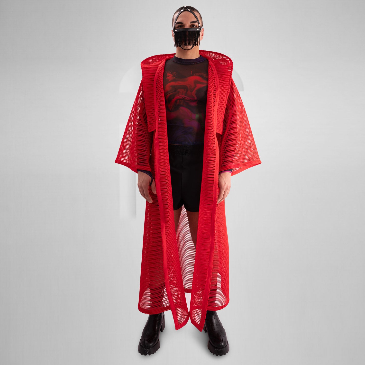 A model wearing a sheer black top with a dramatic red mesh floor-length robe, accessorized with a metallic collar, epitomizing avant-garde fashion and clubwear style, against a neutral gray background.