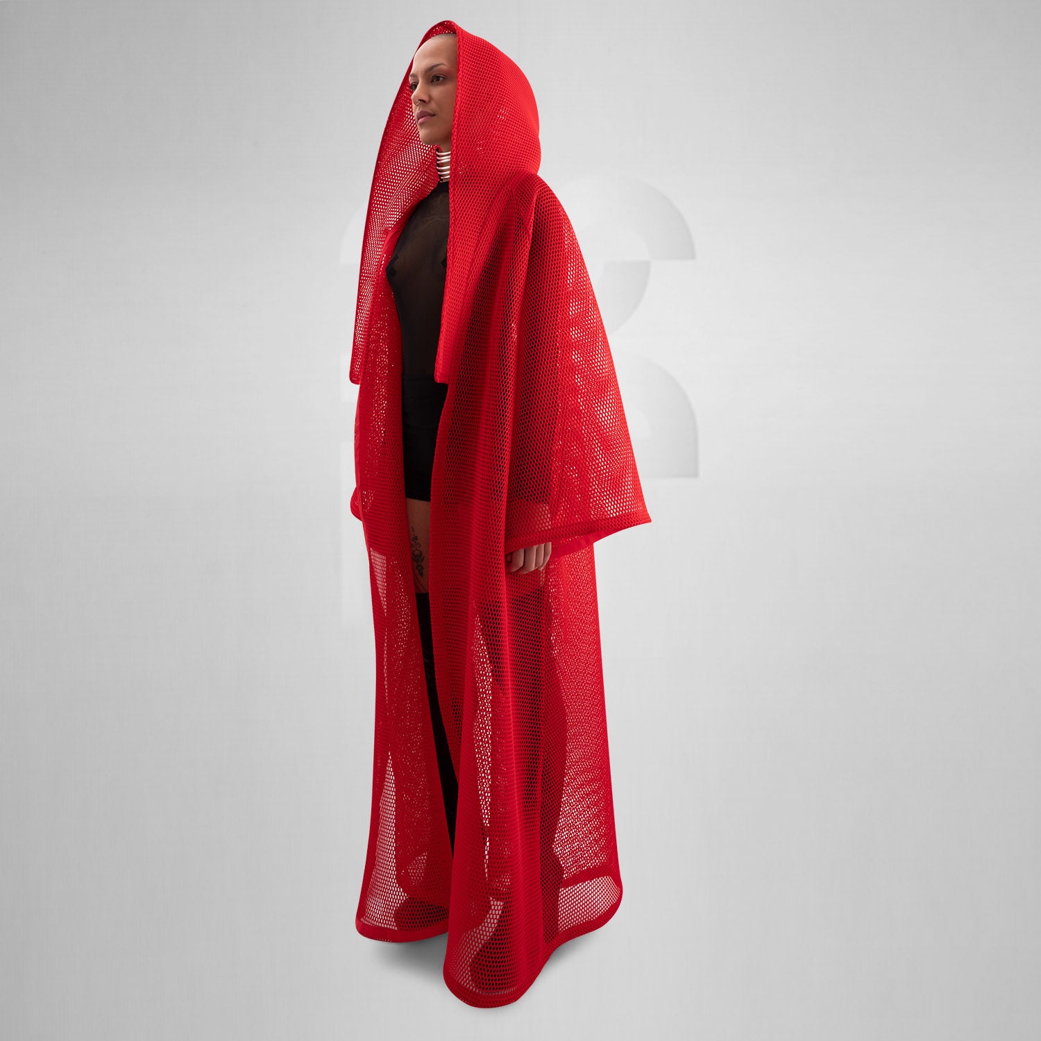 A model wearing a sheer black top with a dramatic red mesh floor-length robe, accessorized with a metallic collar, epitomizing avant-garde fashion and clubwear style, against a neutral gray background.