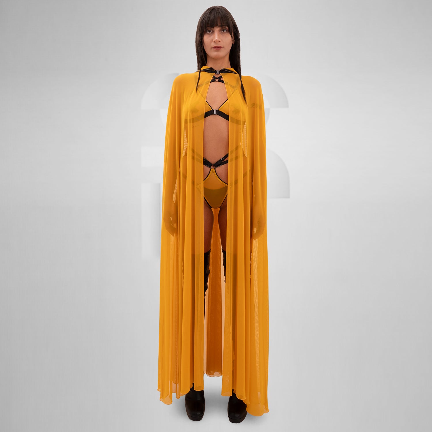 The image features a person wearing a flowing, sheer yellow cape that drapes elegantly to the floor. The cape is lightweight and creates a striking, dramatic silhouette. Underneath, the person is styled in minimal, edgy fashion, with strappy details adding a touch of avant-garde flair. The combination of the vibrant yellow cape and bold styling elements makes for a standout look that’s perfect for fashion-forward occasions.