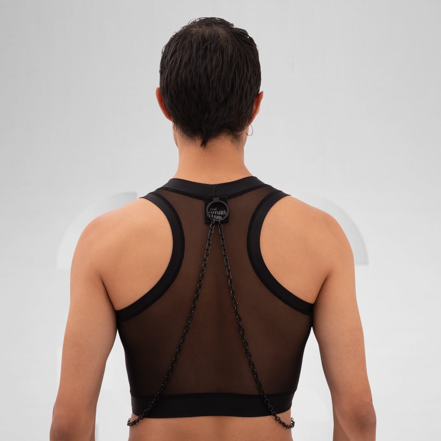 A male model is wearing a black cropped tank top with a sheer mesh fabric covering the torso. The design features a circular metal ring at the center of the chest, with thin black chains extending from the ring to the shoulders and waistband, adding an edgy, modern touch. The background is plain and minimal, emphasizing the bold details of the garment.