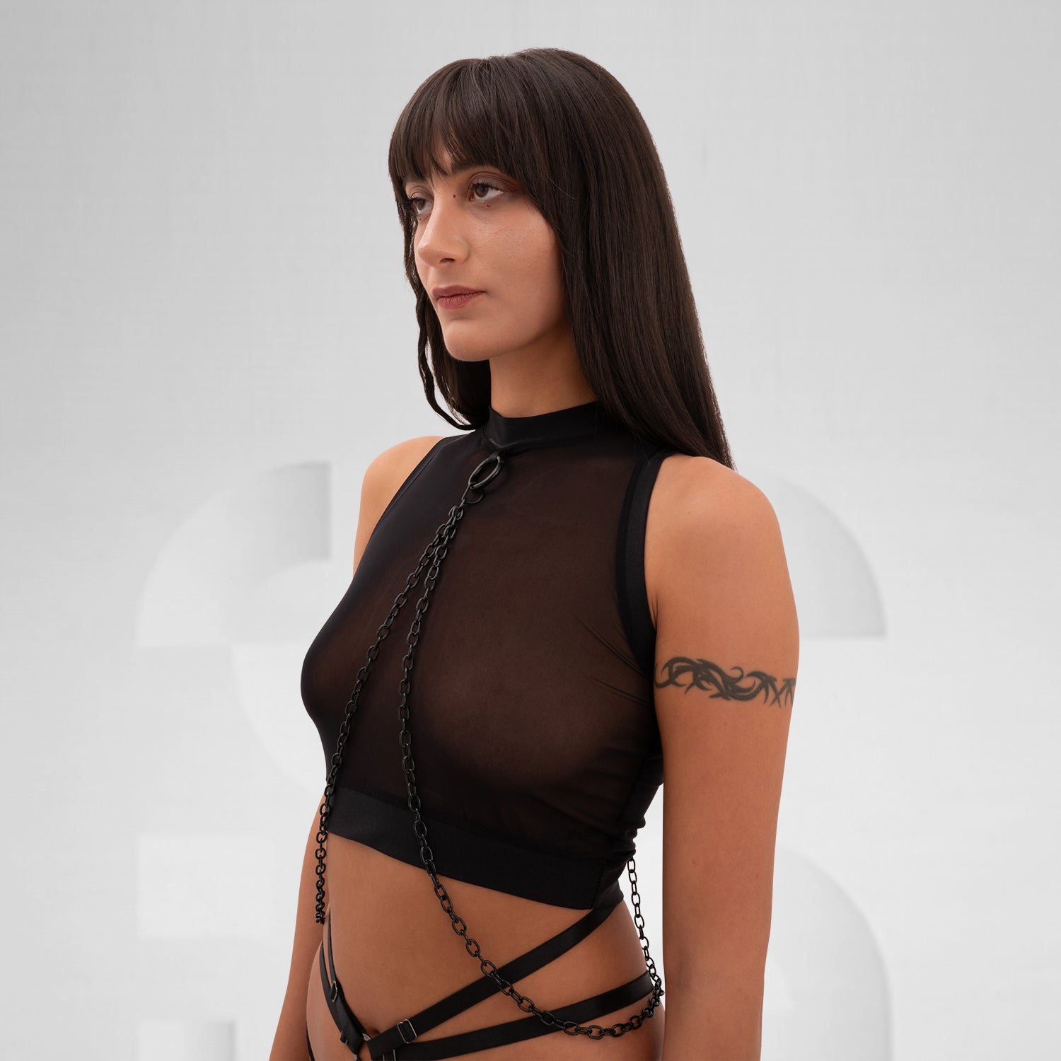 A female model is wearing a black cropped tank top with a sheer mesh fabric covering the torso. The design features a circular metal ring at the center of the chest, with thin black chains extending from the ring to the shoulders and waistband, adding an edgy, modern touch. The background is plain and minimal, emphasizing the bold details of the garment.