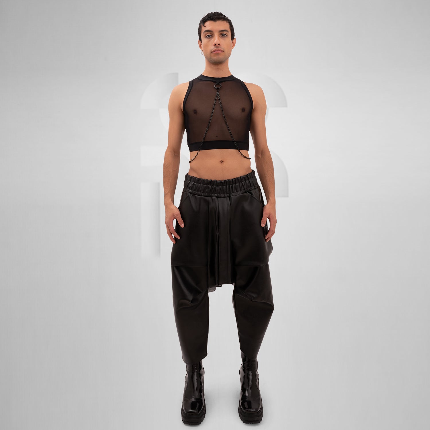 A male model is wearing a black cropped tank top with a sheer mesh fabric covering the torso. The design features a circular metal ring at the center of the chest, with thin black chains extending from the ring to the shoulders and waistband, adding an edgy, modern touch. The background is plain and minimal, emphasizing the bold details of the garment.