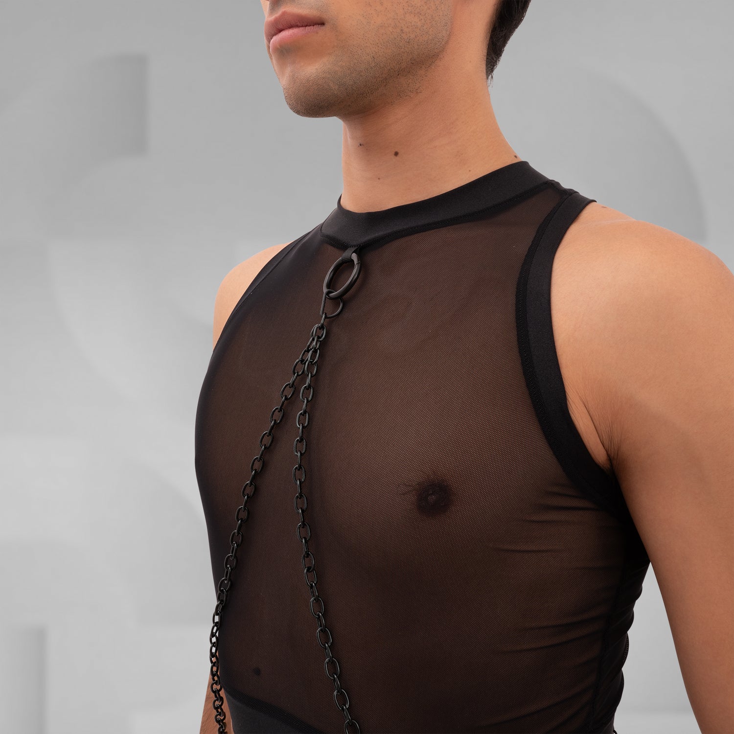 A male model is wearing a black cropped tank top with a sheer mesh fabric covering the torso. The design features a circular metal ring at the center of the chest, with thin black chains extending from the ring to the shoulders and waistband, adding an edgy, modern touch. The background is plain and minimal, emphasizing the bold details of the garment.