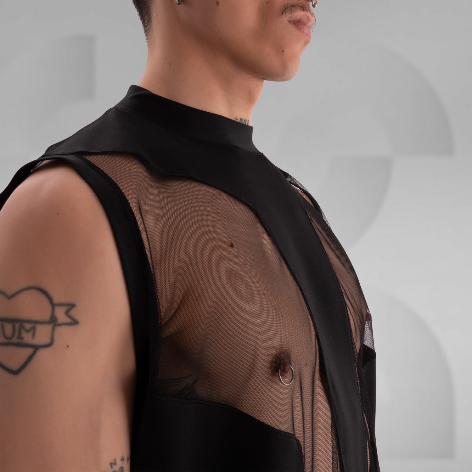 Person wearing a black sleeveless tank top with bold mesh cut-out panels revealing the chest, paired with shiny black vinyl shorts, standing against a neutral background.