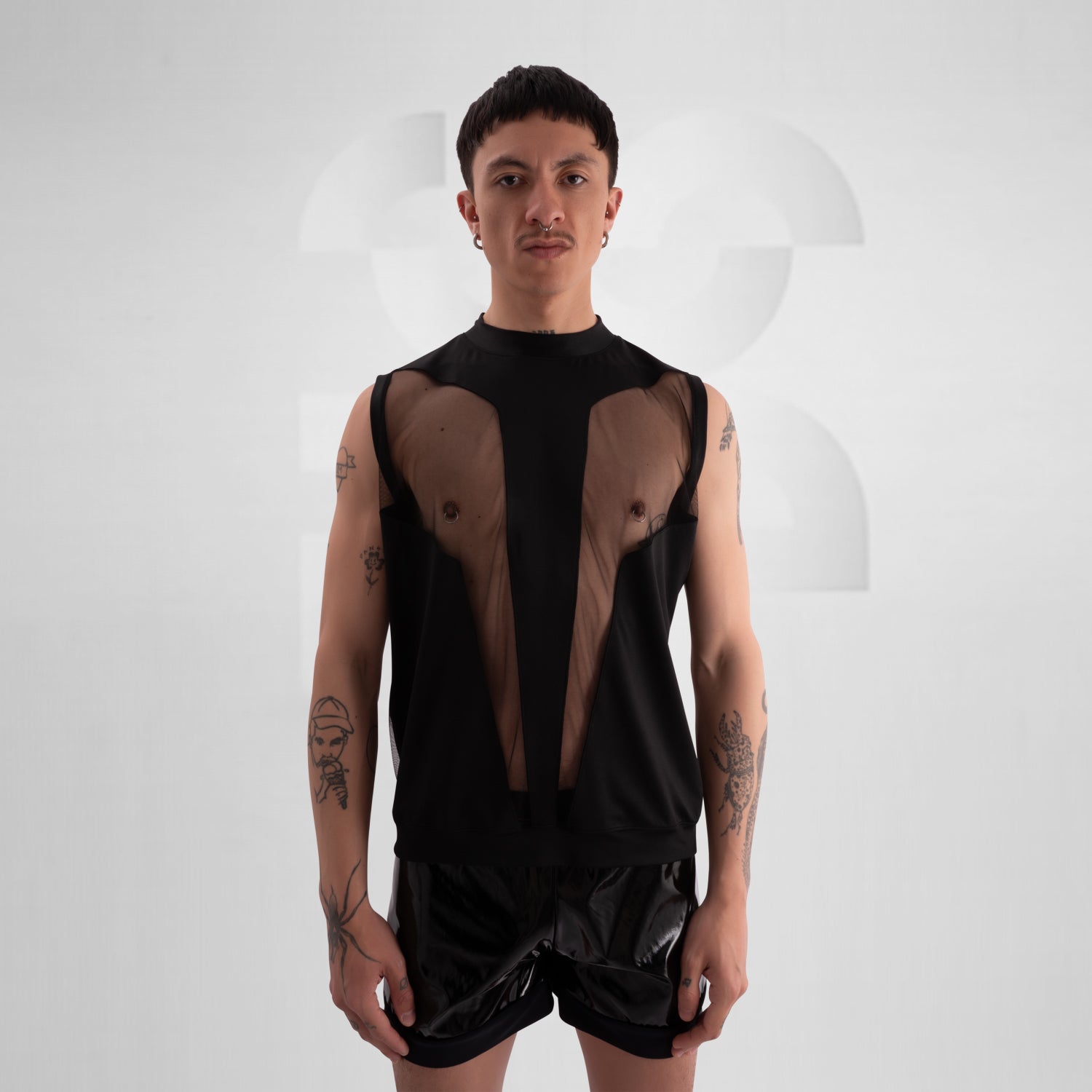 Person wearing a black sleeveless tank top with bold mesh cut-out panels revealing the chest, paired with shiny black vinyl shorts, standing against a neutral background.