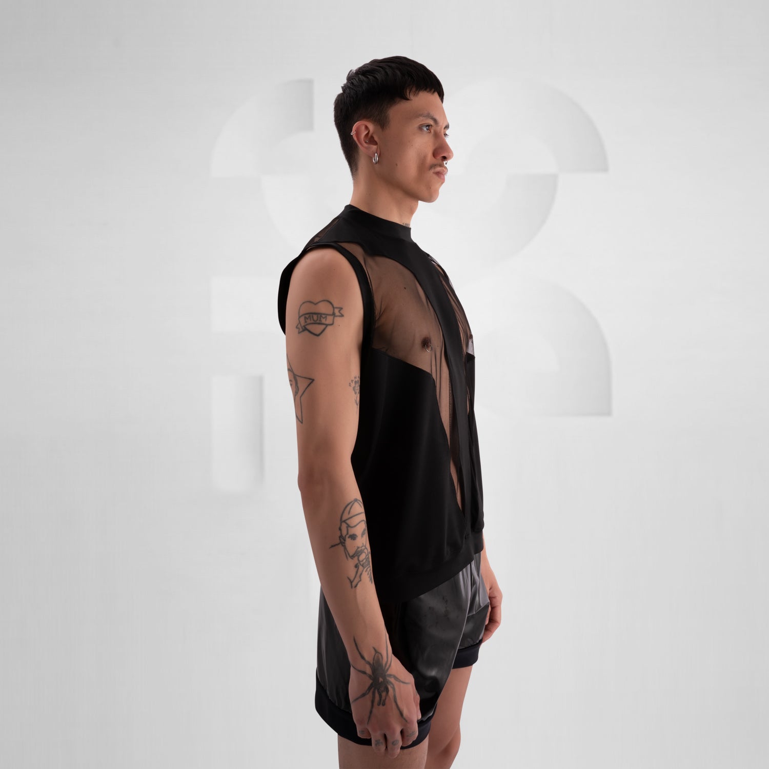 Person wearing a black sleeveless tank top with bold mesh cut-out panels revealing the chest, paired with shiny black vinyl shorts, standing against a neutral background.