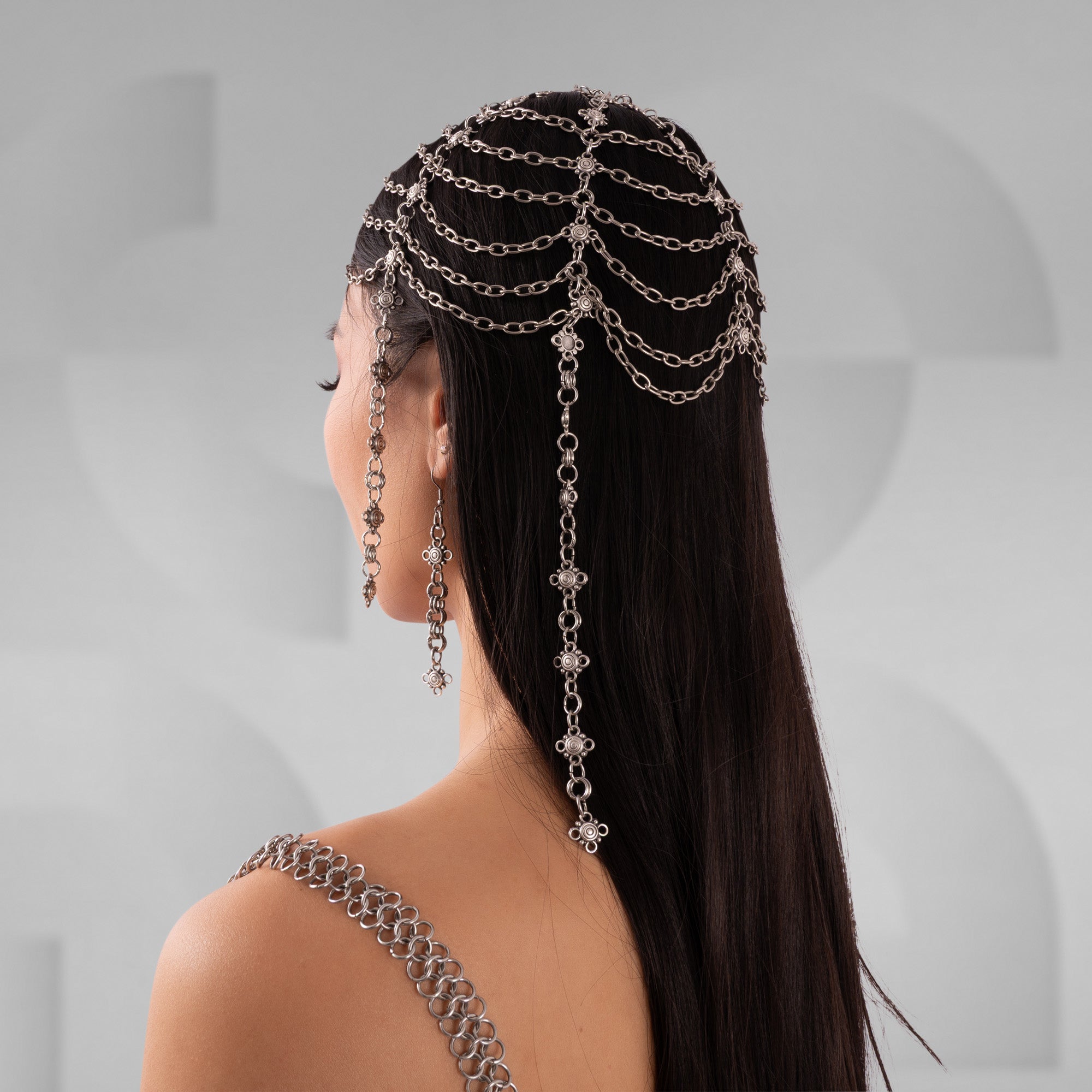 Woman with long dark hair wearing a decorative chain headpiece against a gray background