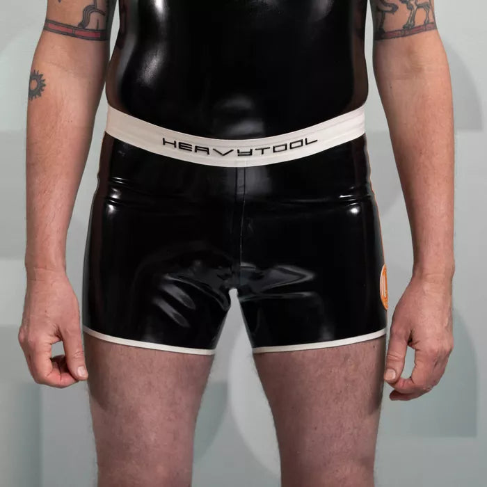 The image features sleek black shorts from HeavyTool Gear, designed for a bold and eye-catching look. The shiny latex material hugs the body, creating a striking silhouette, while the contrasting waistband adds a stylish touch. Perfect for both clubwear and performance, these shorts exude confidence and a unique sense of style.