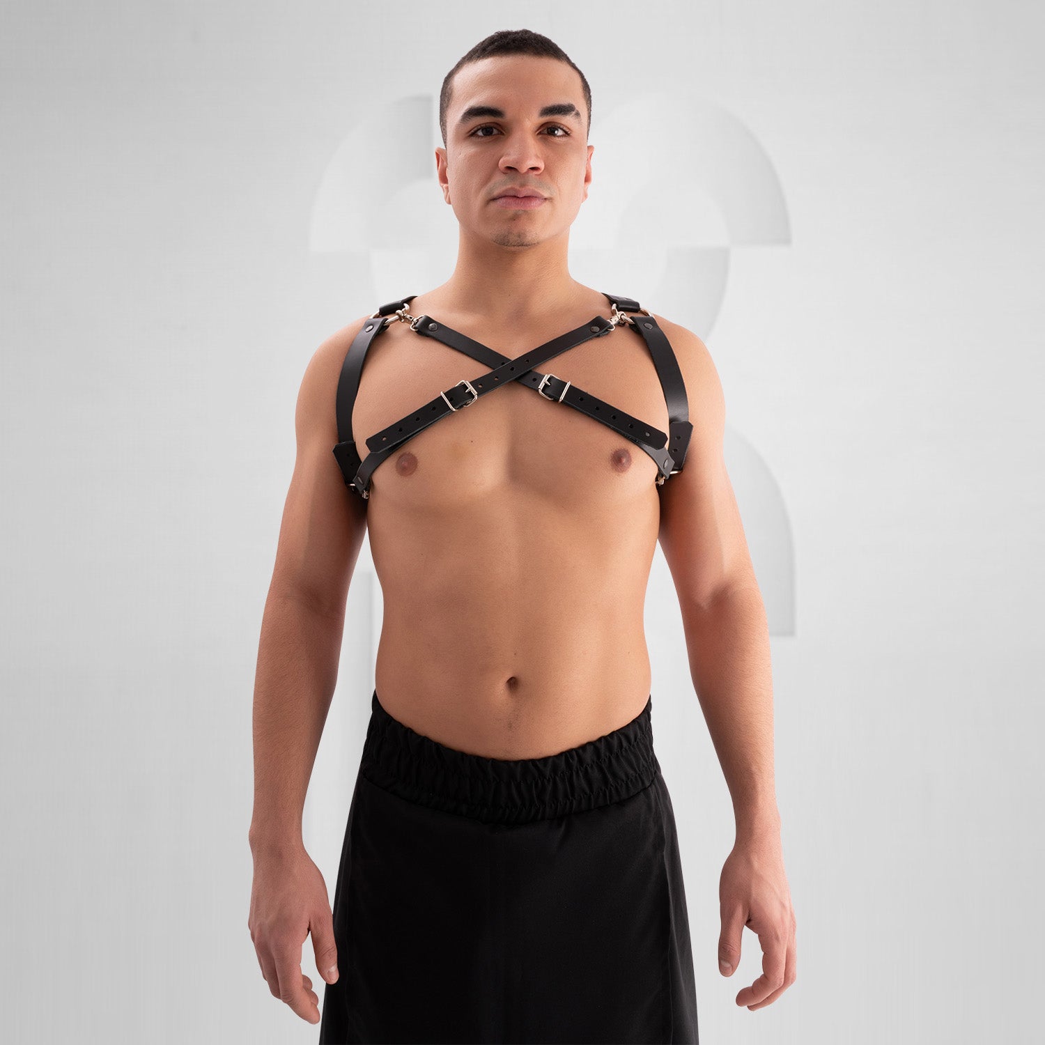 Male model wearing a black leather harness with adjustable buckle straps, featuring a bold crisscross design across the chest. Styled with a black skirt, this avant-garde look is perfect for festival, fetish, and clubwear fashion.