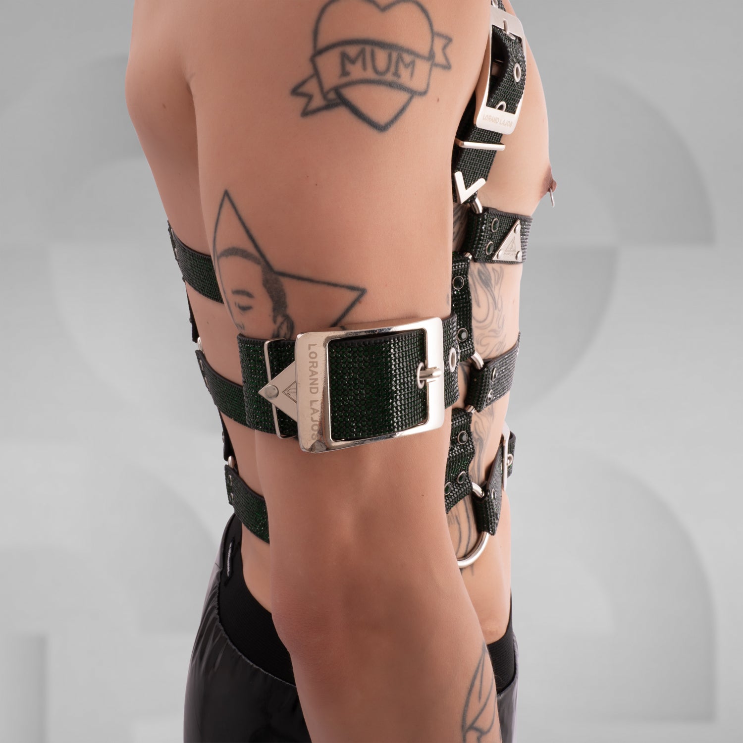 The image displays a close-up of a person's arm wearing a bold, rgreen strap with sparkling embellishments and metallic hardware, including a triangular and circular logo plate. The accessory creates a striking, edgy style statement.