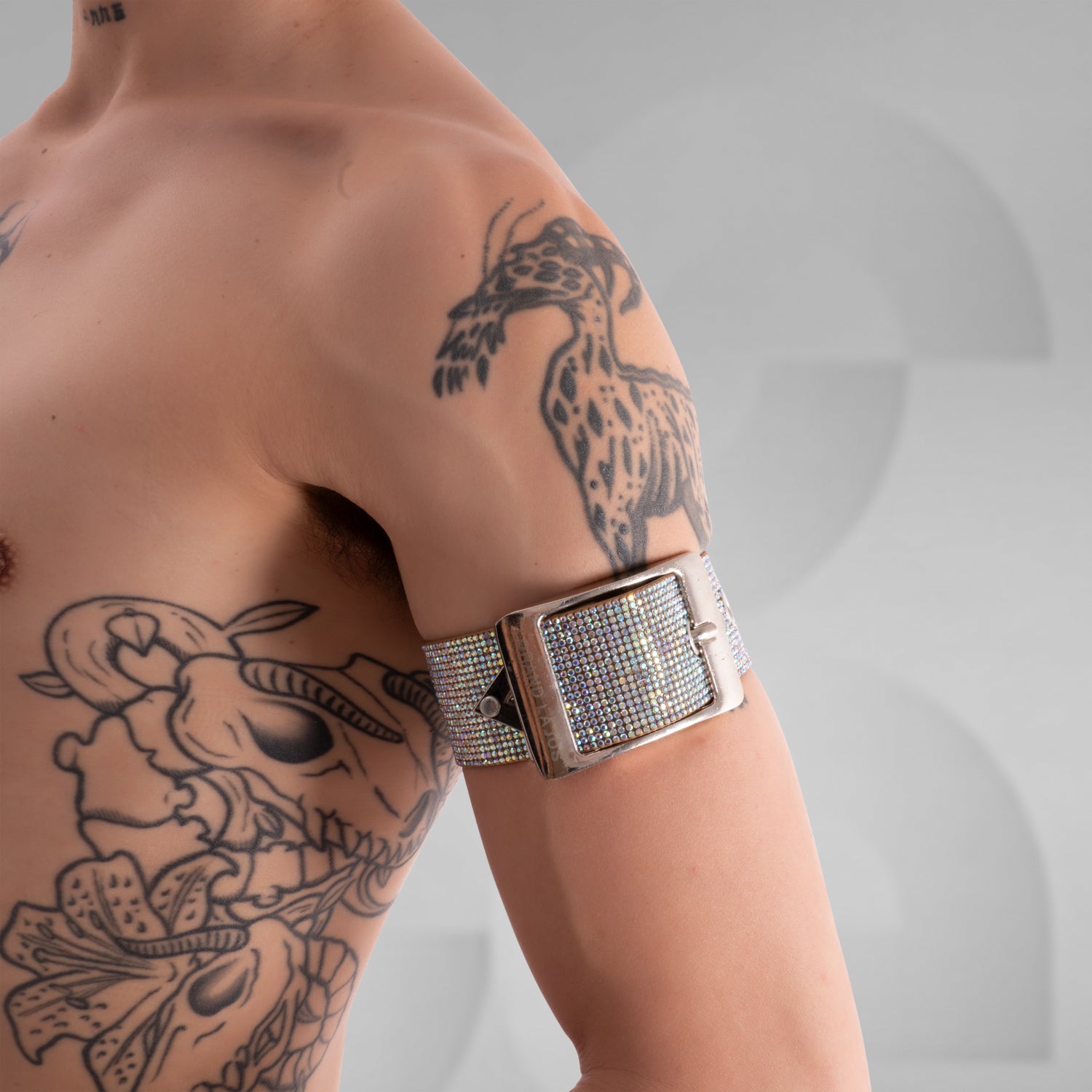 The image displays a close-up of a person's arm wearing a bold, white strap with sparkling embellishments and metallic hardware, including a triangular and circular logo plate. The accessory creates a striking, edgy style statement.