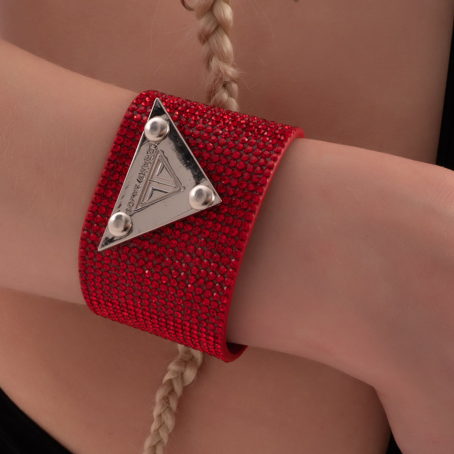 Close-up of a model wearing a striking red crystal-studded bracelet with a silver triangular emblem from Lorand Lajos, styled with a black latex cut-out bodysuit and long braided hair – edgy, glamorous, and festival-ready.