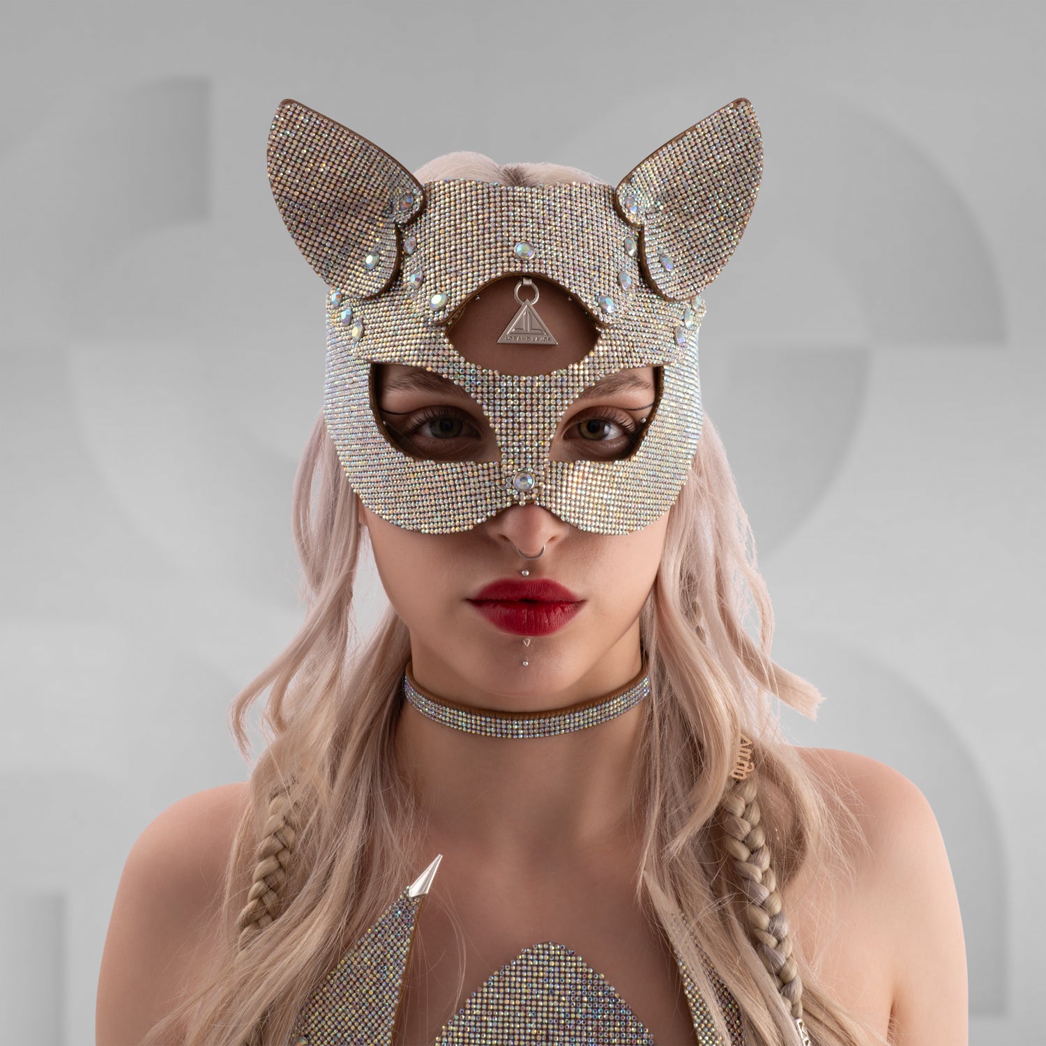 Woman wearing a sparkling rhinestone cat mask with pointed ears, paired with a matching crystal choker and shimmering outfit; styled with platinum blonde hair in braids and bold red lipstick, set against a minimalist grey background.