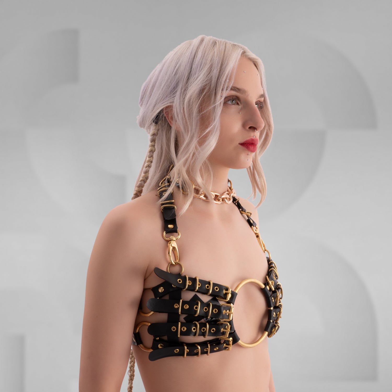 Side view of a model wearing a bold, avant-garde leather harness bra with multiple black leather straps, gold metal buckles, and a large central gold ring; styled with a chunky gold chain necklace and platinum blonde braided hair, set against a neutral geometric background.