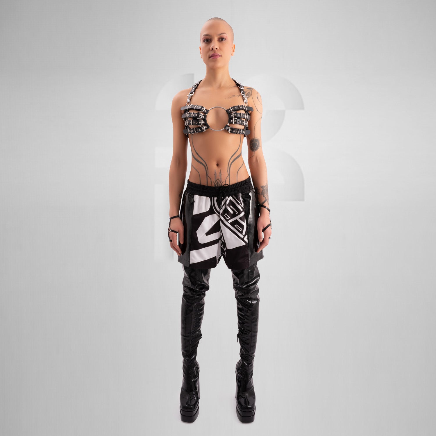 Striking avant-garde rubber bra composed of multiple black straps with silver metal buckles, connected by a large central O-ring. The harness-style design creates a bold, industrial look, worn by a confident model with a shaved head and visible tattoos, standing against a minimalist grey background.
