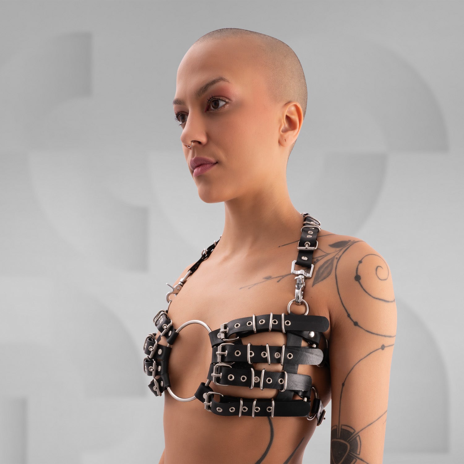 Striking avant-garde rubber bra composed of multiple black straps with silver metal buckles, connected by a large central O-ring. The harness-style design creates a bold, industrial look, worn by a confident model with a shaved head and visible tattoos, standing against a minimalist grey background.