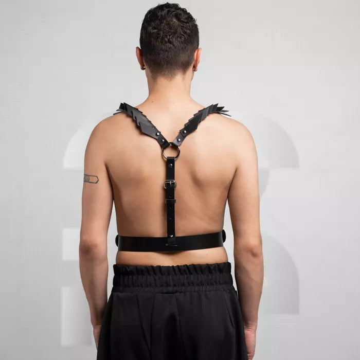 is wearing a bold black leather harness with decorative details on the shoulders, paired with loose black pants. The look is edgy and minimalist, set against a neutral background that highlights the outfit's unique design.