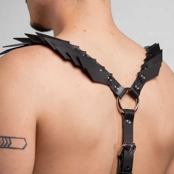 is wearing a bold black leather harness with decorative details on the shoulders, paired with loose black pants. The look is edgy and minimalist, set against a neutral background that highlights the outfit's unique design.