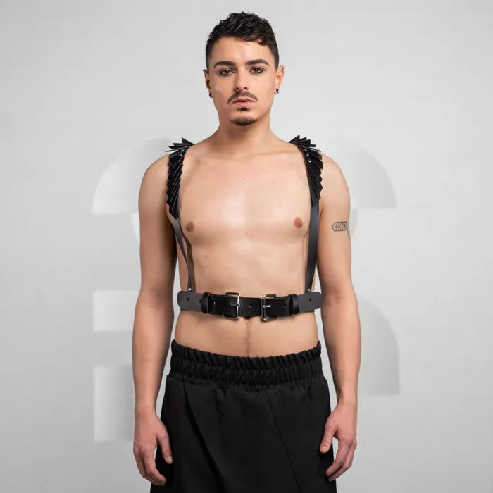 is wearing a bold black leather harness with decorative details on the shoulders, paired with loose black pants. The look is edgy and minimalist, set against a neutral background that highlights the outfit's unique design.