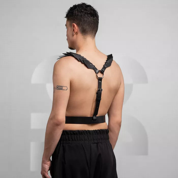 is wearing a bold black leather harness with decorative details on the shoulders, paired with loose black pants. The look is edgy and minimalist, set against a neutral background that highlights the outfit's unique design.