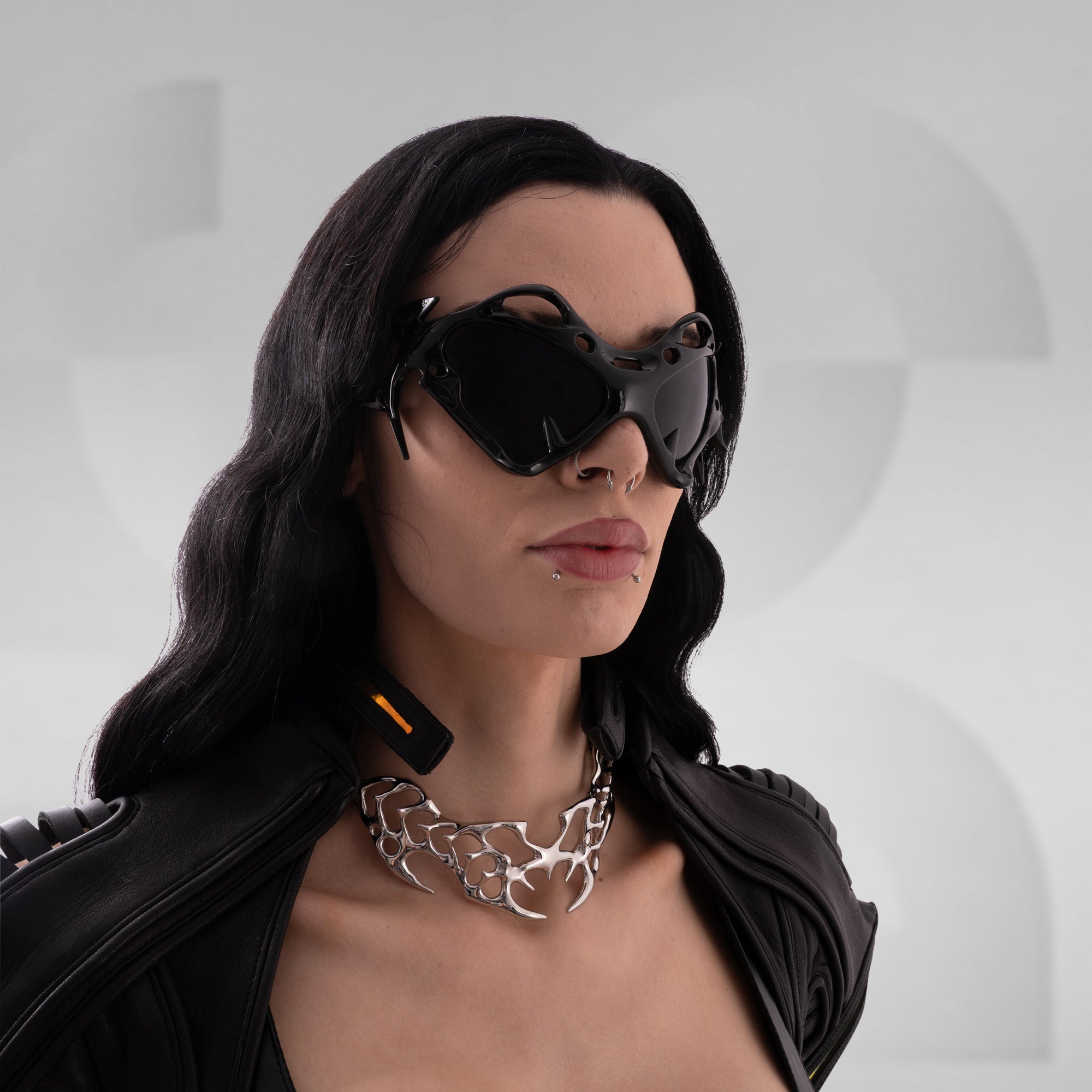 Woman wearing futuristic sunglasses and a black leather jacket with a silver necklace on a gray background