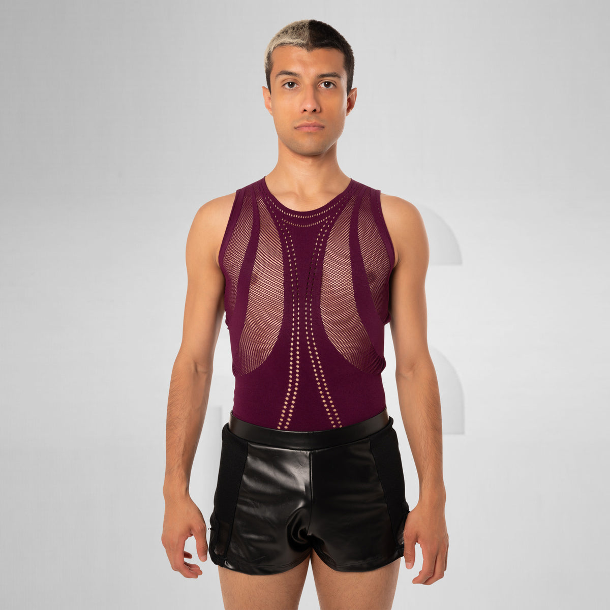 A male model wearing a sleeveless burgundy mesh athletic tank top with structured underbust support and black shorts, standing against a light gray studio background.