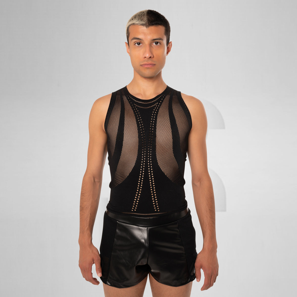 A male model wearing a black sleeveless mesh bodysuit with gold-tone stud detailing and black shorts, standing against a light gray studio background.