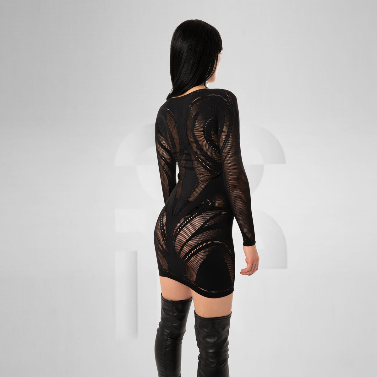 A model shown from the back wearing a black sheer long-sleeve mini dress with geometric cutout patterns and black thigh-high boots, set against a light gray studio background.