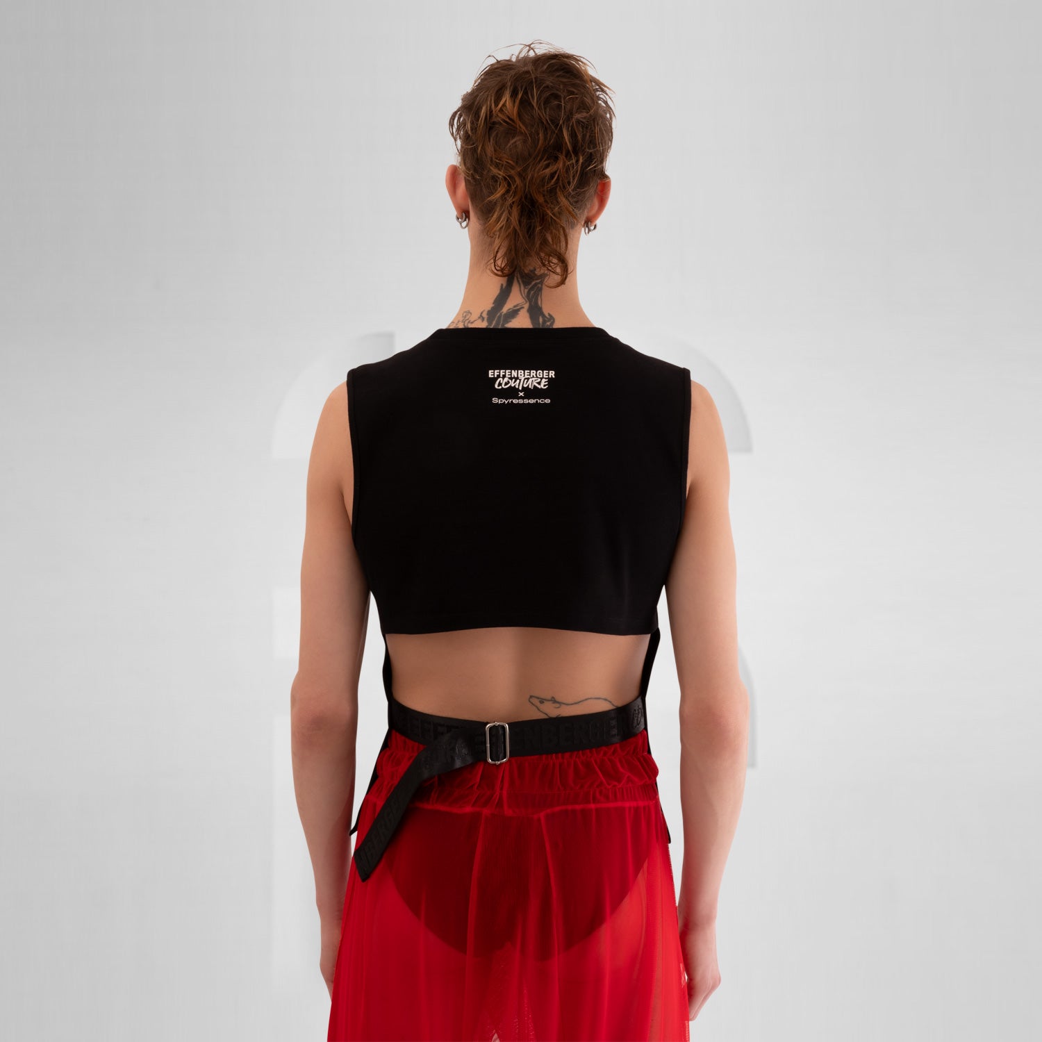 The image features a person wearing a sleeveless black shirt with a striking graphic print on the front, paired with red shorts. The background is a neutral gray, emphasizing the bold design of the shirt. This outfit combines edgy fashion with a modern, artistic flair.