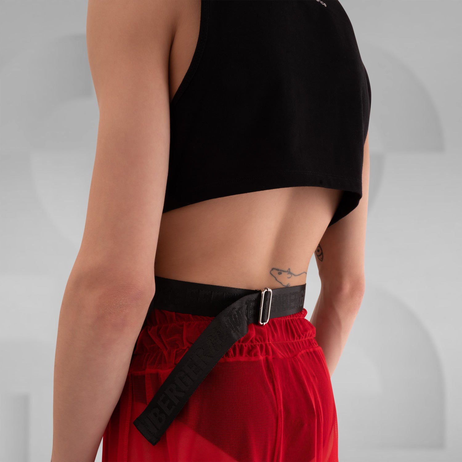 The image features a person wearing a sleeveless black shirt with a striking graphic print on the front, paired with red shorts. The background is a neutral gray, emphasizing the bold design of the shirt. This outfit combines edgy fashion with a modern, artistic flair.