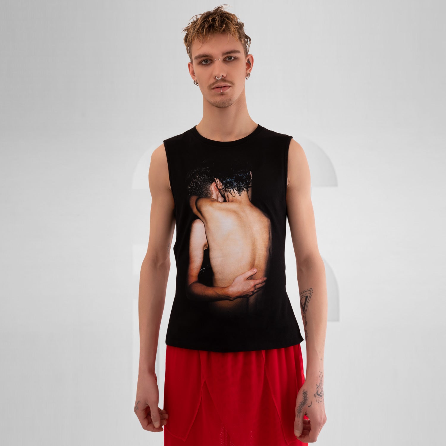 The image features a person wearing a sleeveless black shirt with a striking graphic print on the front, paired with red shorts. The background is a neutral gray, emphasizing the bold design of the shirt. This outfit combines edgy fashion with a modern, artistic flair.