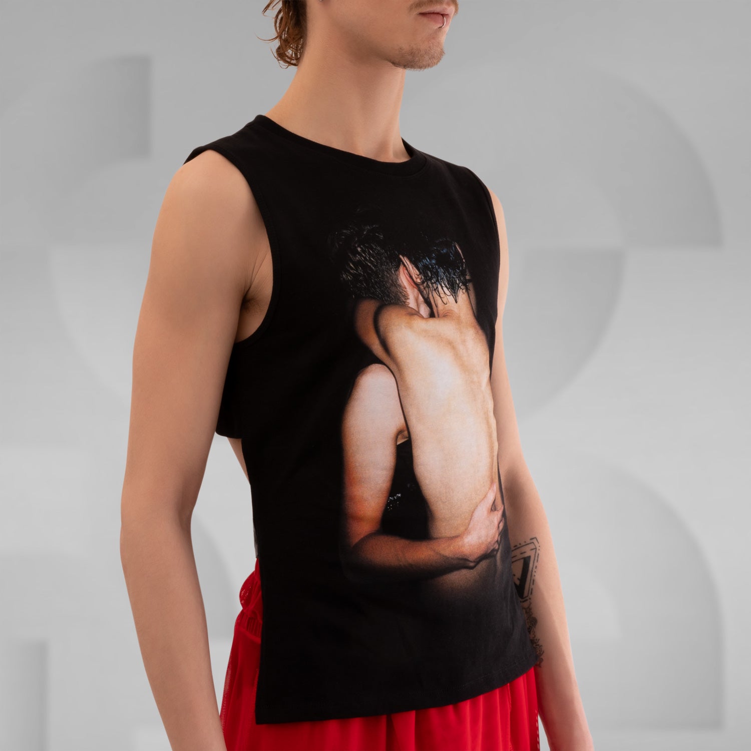 The image features a person wearing a sleeveless black shirt with a striking graphic print on the front, paired with red shorts. The background is a neutral gray, emphasizing the bold design of the shirt. This outfit combines edgy fashion with a modern, artistic flair.