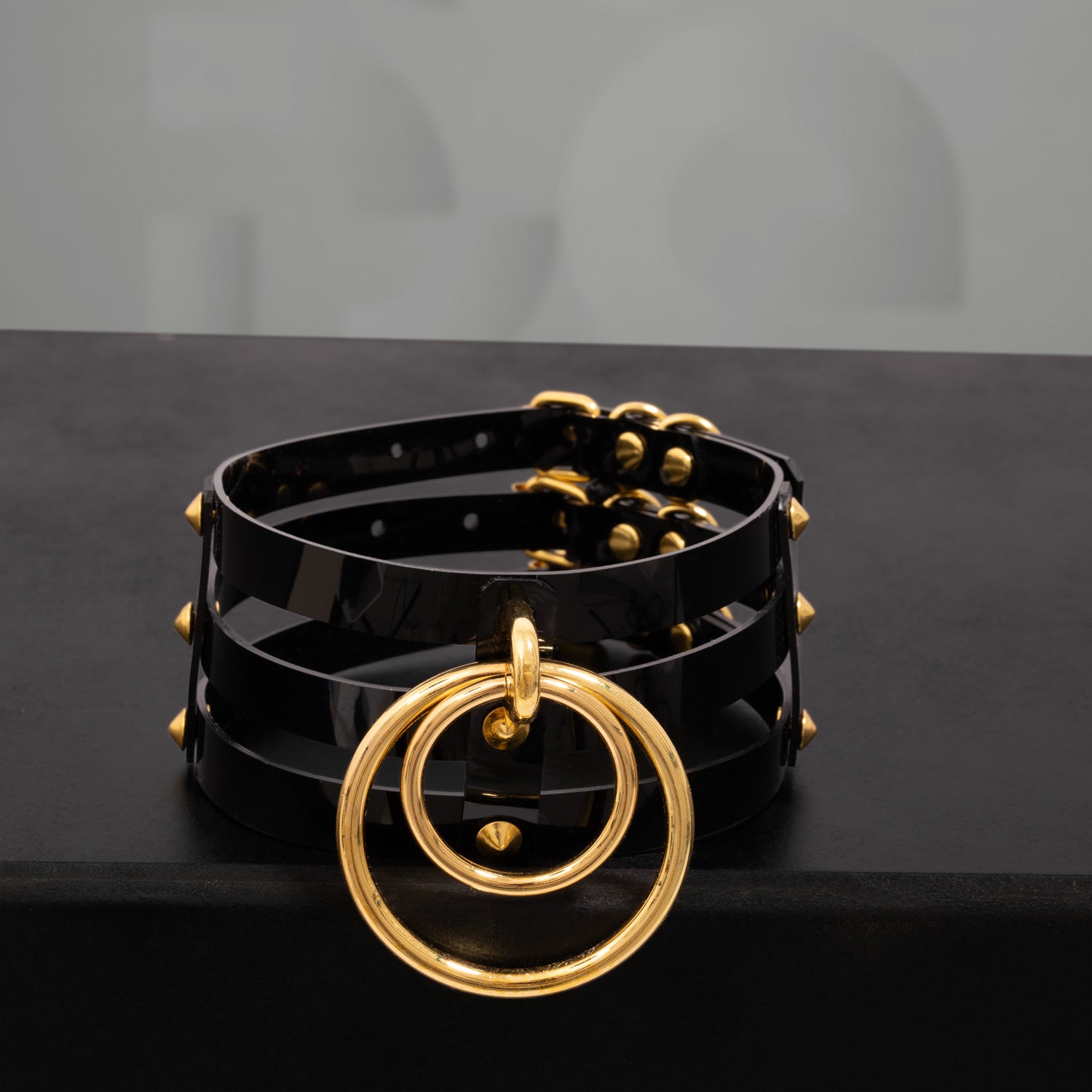 This image showcases a striking black PVC choker featuring three sleek stripes and adorned with metallic spikes. The design includes two bold rings at the front, adding an edgy yet elegant touch. The high-gloss finish of the PVC enhances its modern aesthetic, making it a perfect accessory for both nightlife and everyday outfits. This choker is designed to make a statement while blending a sense of daring style with sophistication.