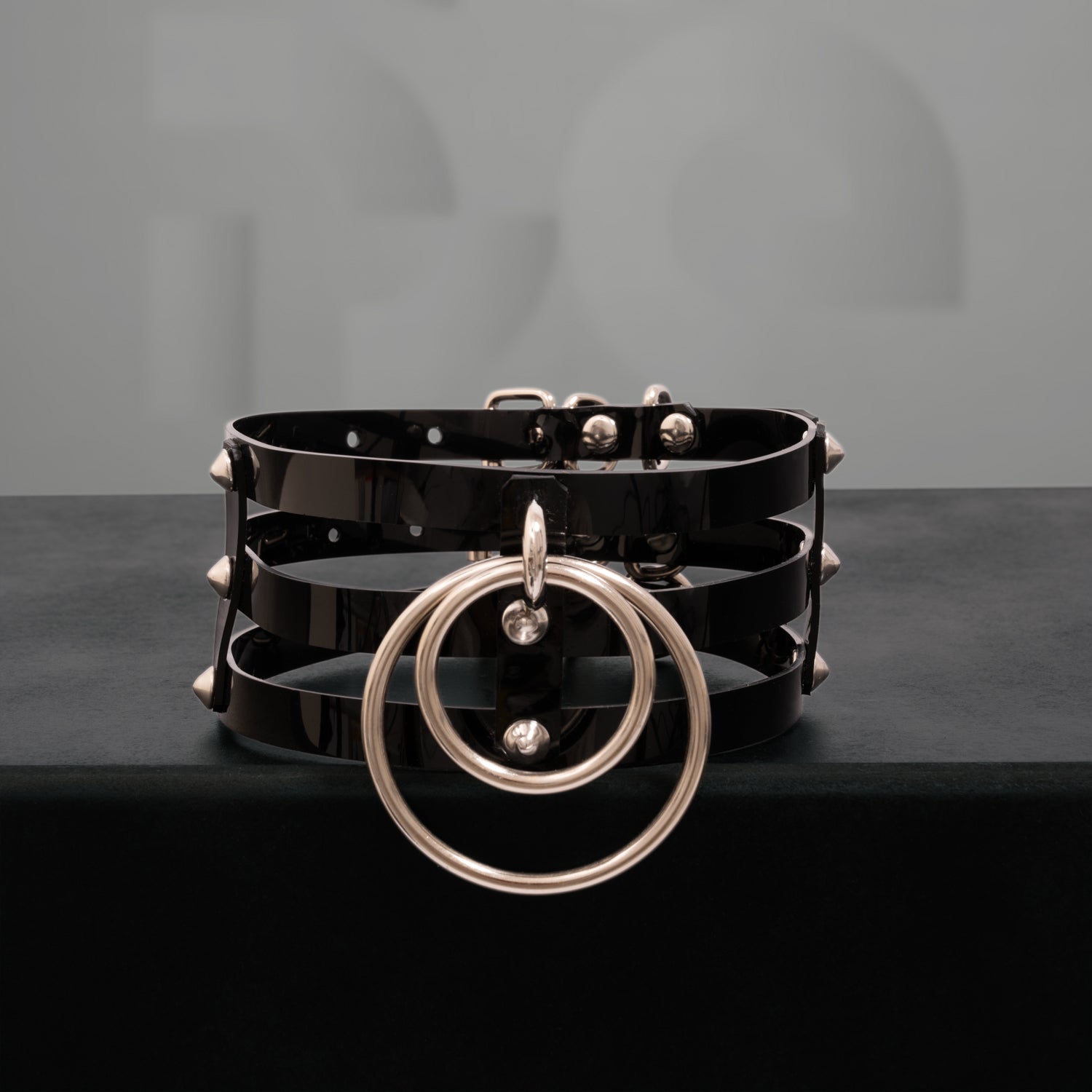 This image showcases a striking black PVC choker featuring three sleek stripes and adorned with metallic spikes. The design includes two bold rings at the front, adding an edgy yet elegant touch. The high-gloss finish of the PVC enhances its modern aesthetic, making it a perfect accessory for both nightlife and everyday outfits. This choker is designed to make a statement while blending a sense of daring style with sophistication.