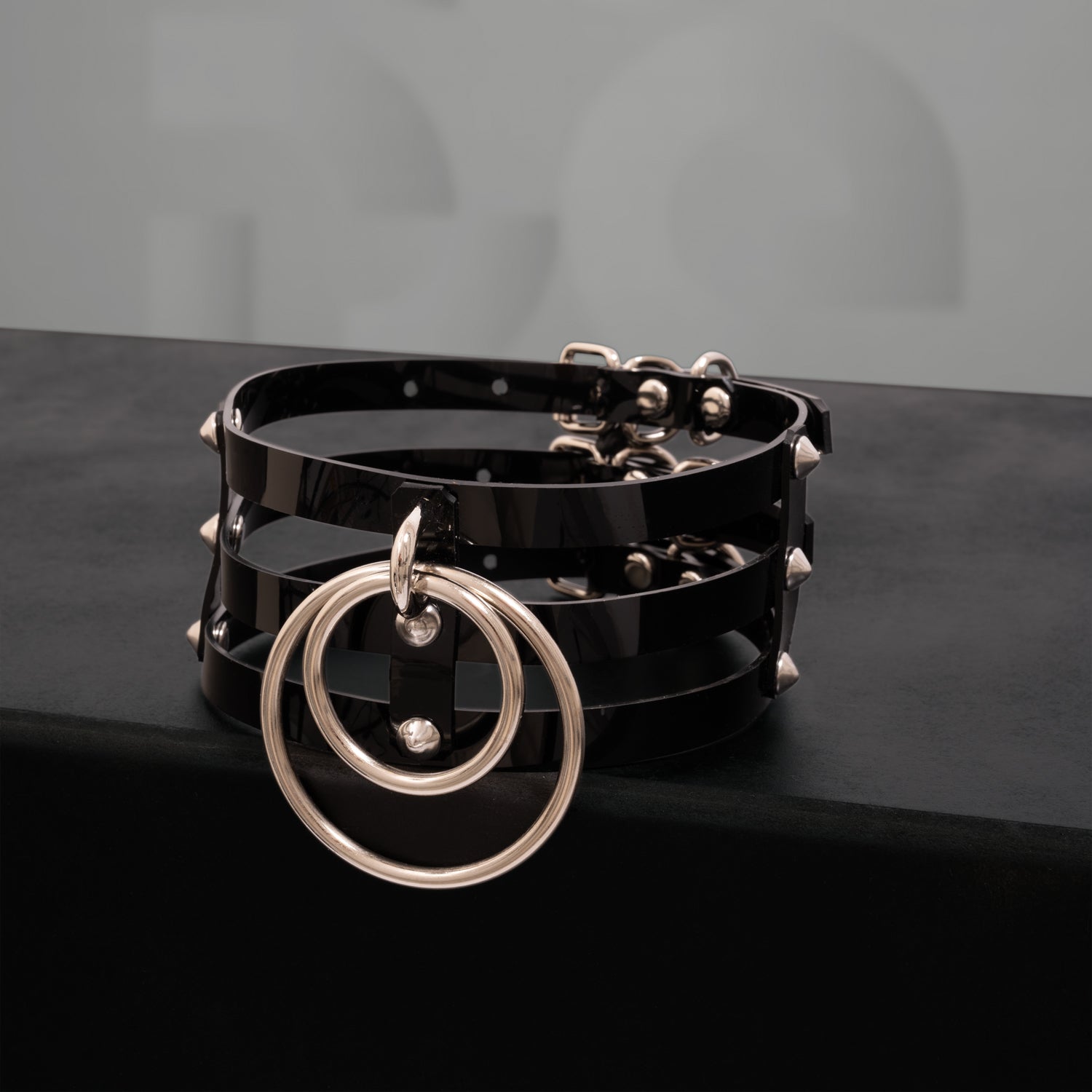 This image showcases a striking black PVC choker featuring three sleek stripes and adorned with metallic spikes. The design includes two bold rings at the front, adding an edgy yet elegant touch. The high-gloss finish of the PVC enhances its modern aesthetic, making it a perfect accessory for both nightlife and everyday outfits. This choker is designed to make a statement while blending a sense of daring style with sophistication.