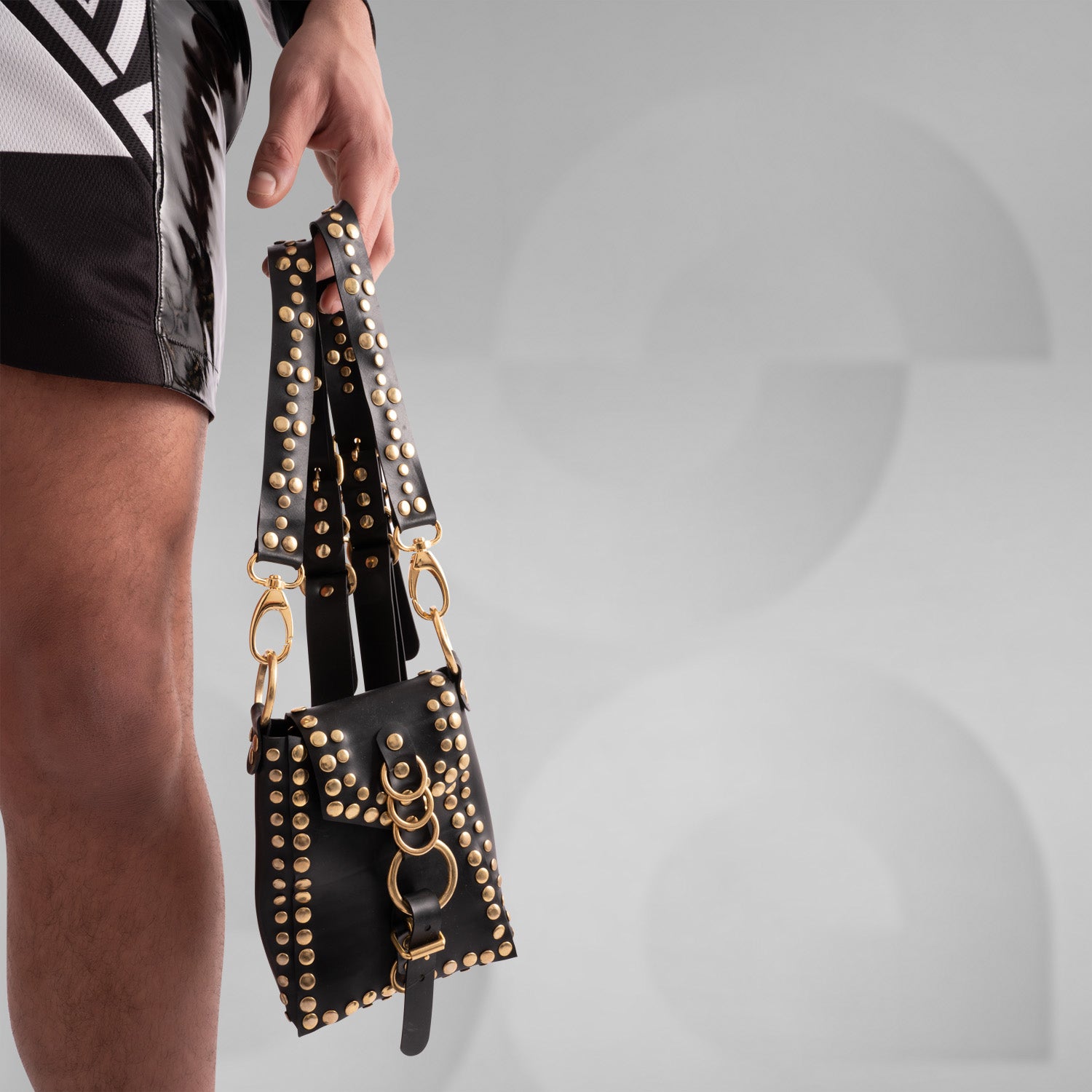 Person wearing a black studded leather harness bag with silver hardware and rings, styled over a sheer mesh top featuring vibrant red and purple abstract artwork. The edgy, fashion-forward look is completed with holographic geometric-patterned pants, captured from behind against a minimalist grey background.