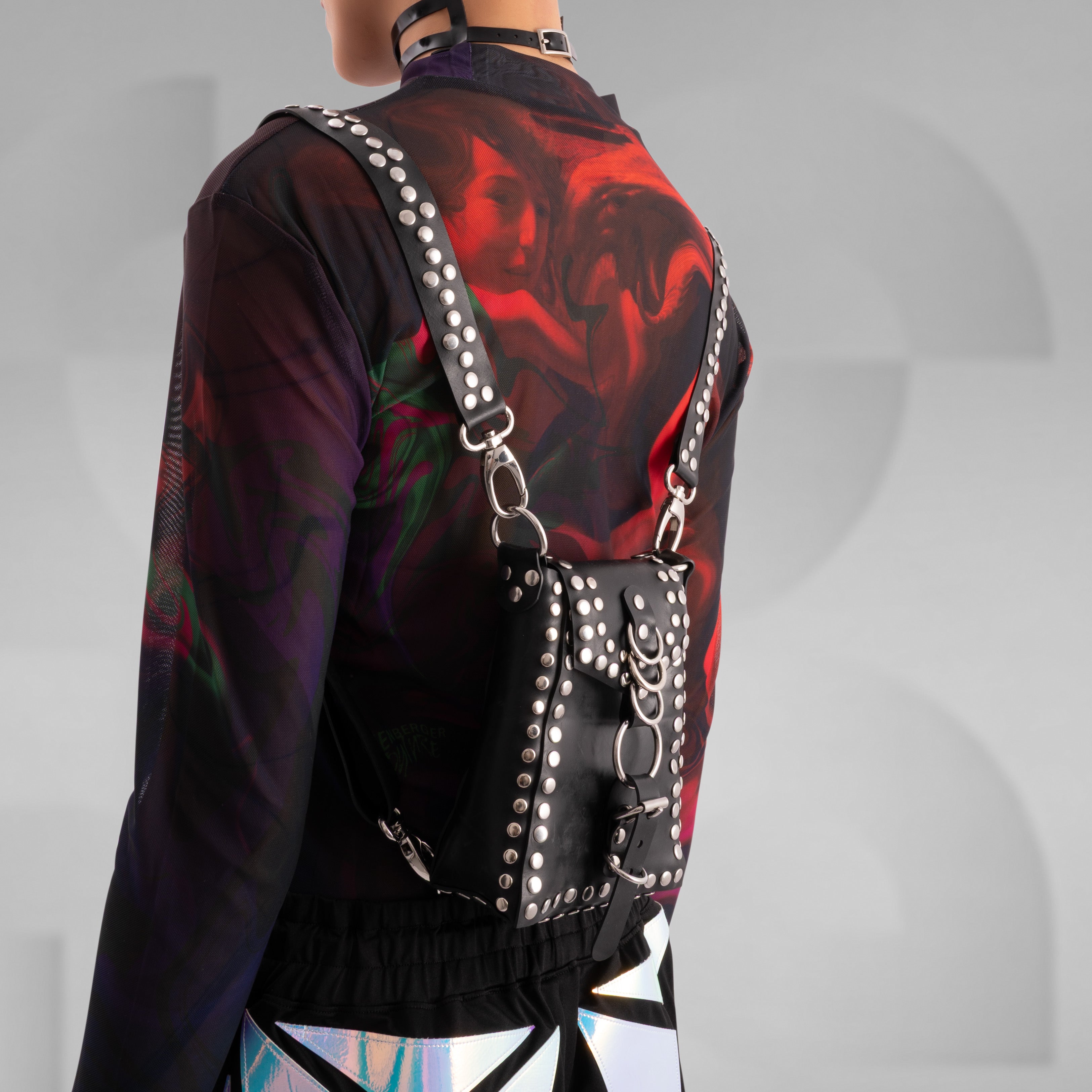 Person wearing a black studded leather harness bag with silver hardware and rings, styled over a sheer mesh top featuring vibrant red and purple abstract artwork. The edgy, fashion-forward look is completed with holographic geometric-patterned pants, captured from behind against a minimalist grey background.