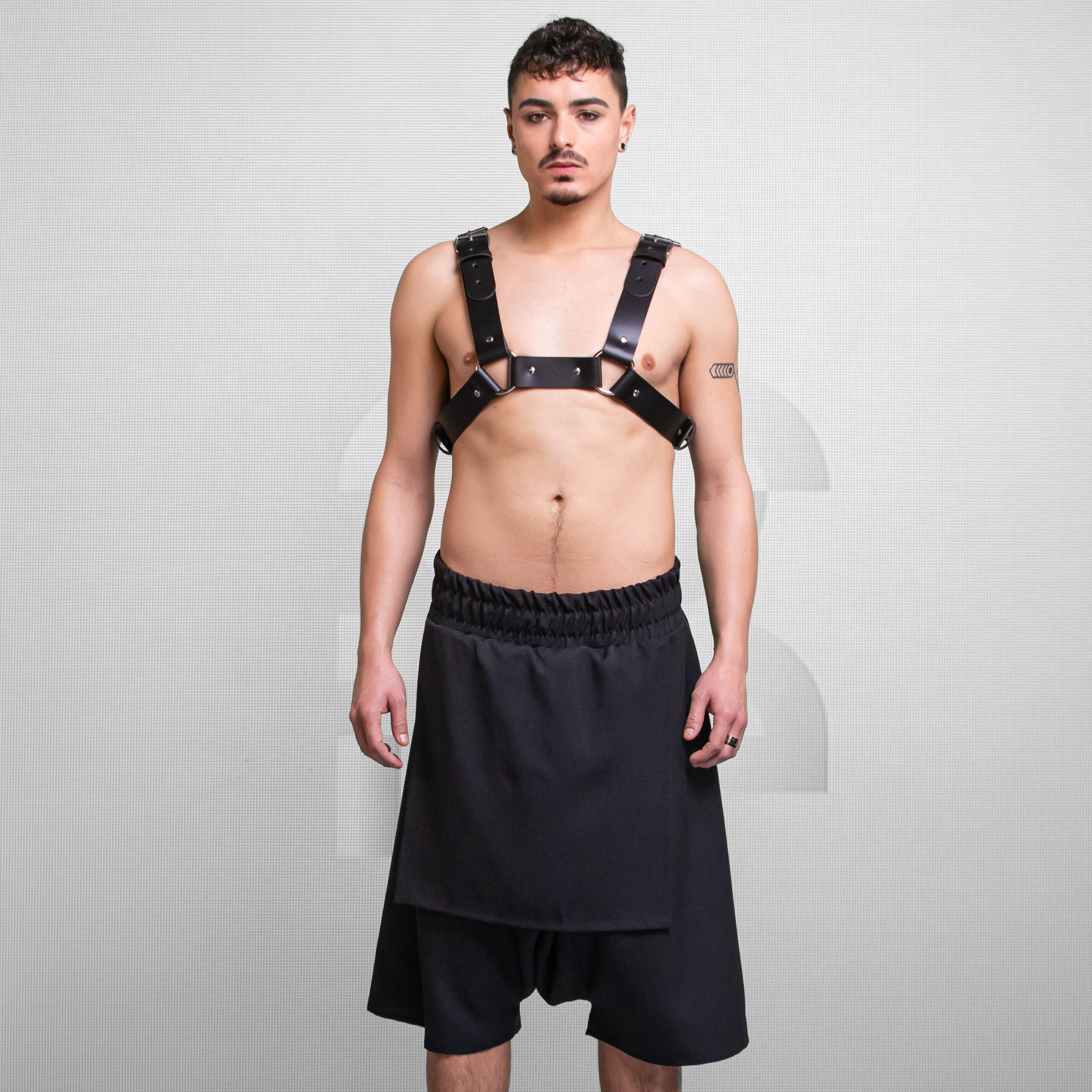 striking black leather harness paired with a flowy black skirt. The harness features bold straps and metallic accents, creating an edgy aesthetic. The look is complemented by the neutral background, emphasizing the outfit's unique combination of strength and style.