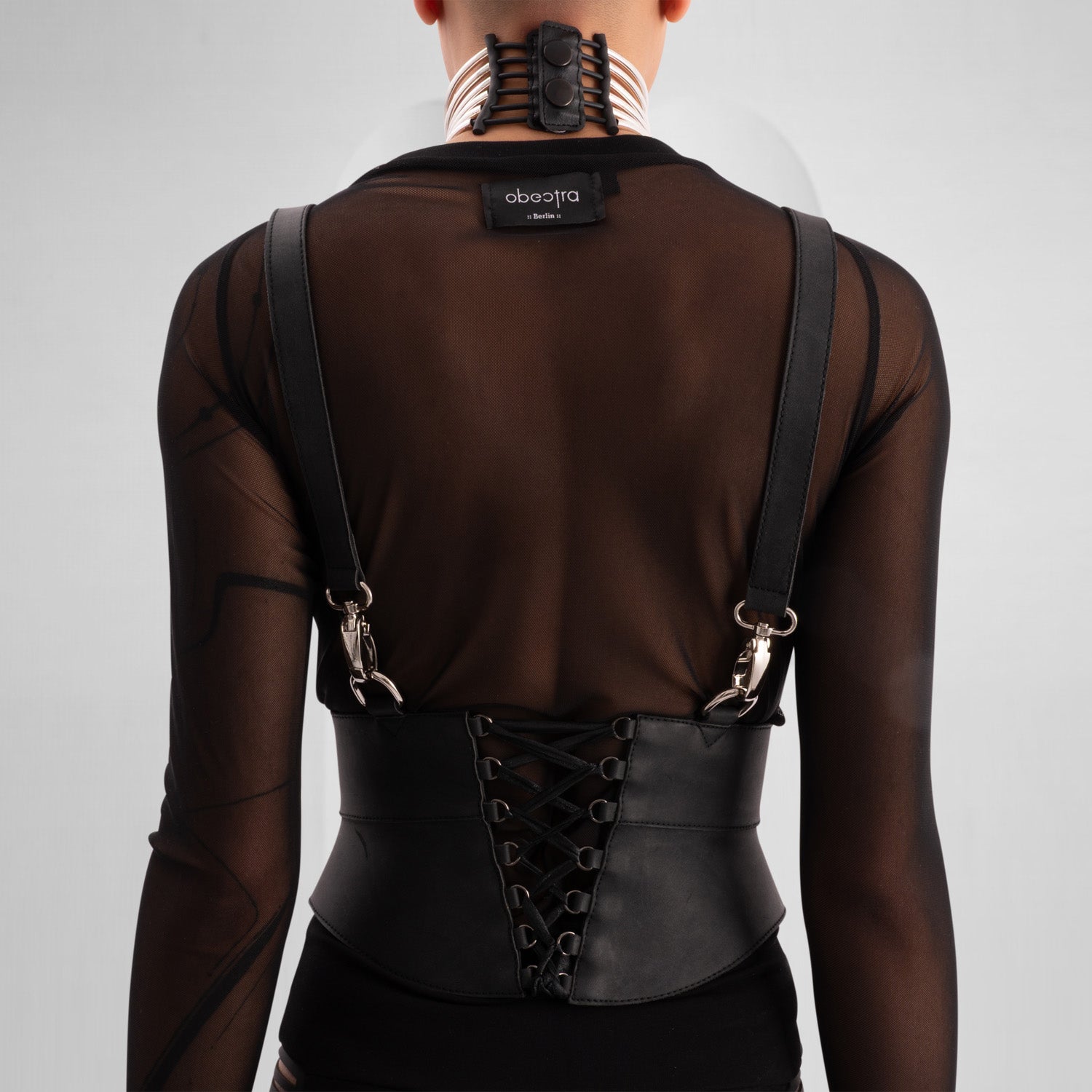 A close-up of a model wearing a sheer black top with decorative straps, featuring a unique leather harness belt and a metallic choker collar, showcasing a bold and edgy fashion statement against a neutral gray background.