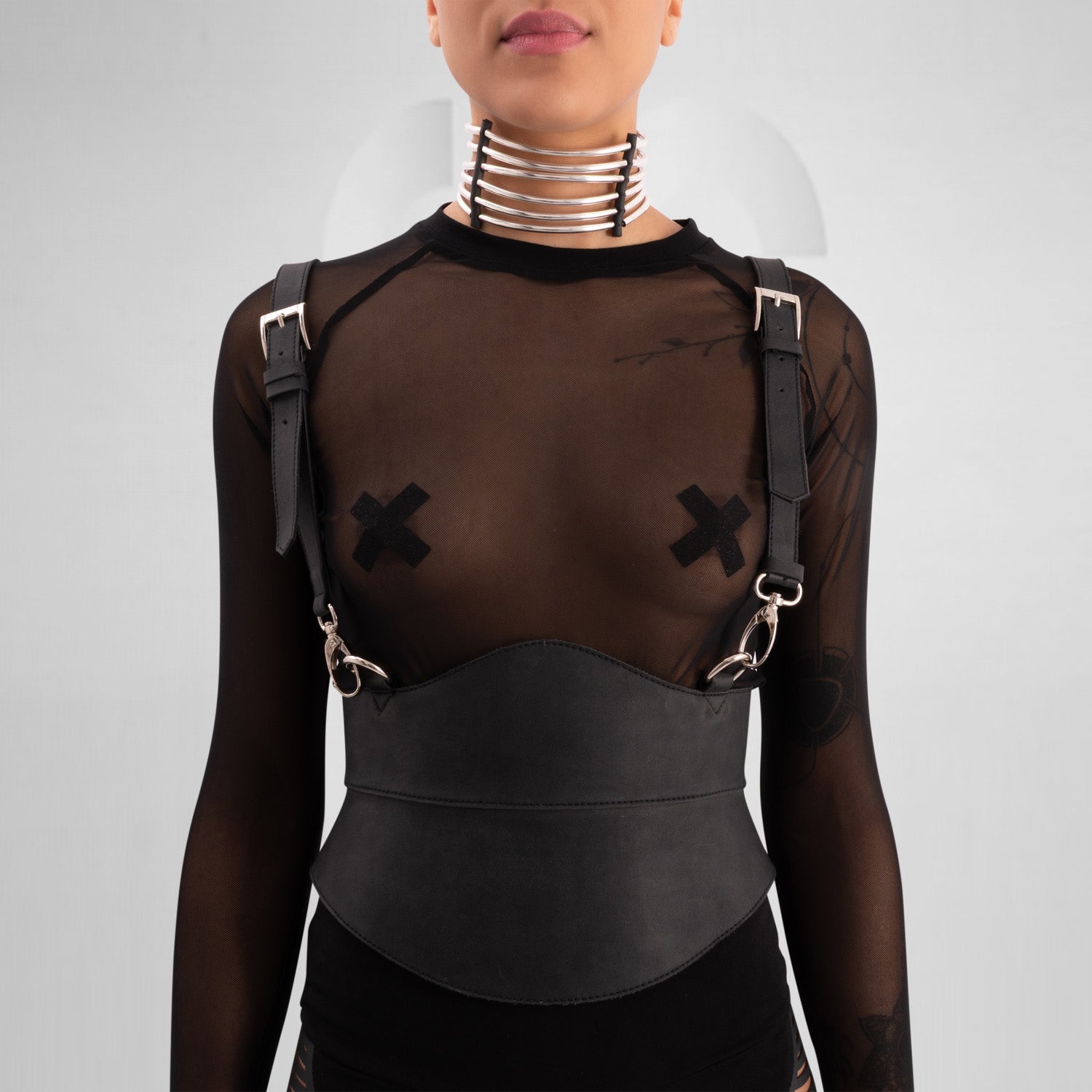 A close-up of a model wearing a sheer black top with decorative straps, featuring a unique leather harness belt and a metallic choker collar, showcasing a bold and edgy fashion statement against a neutral gray background.