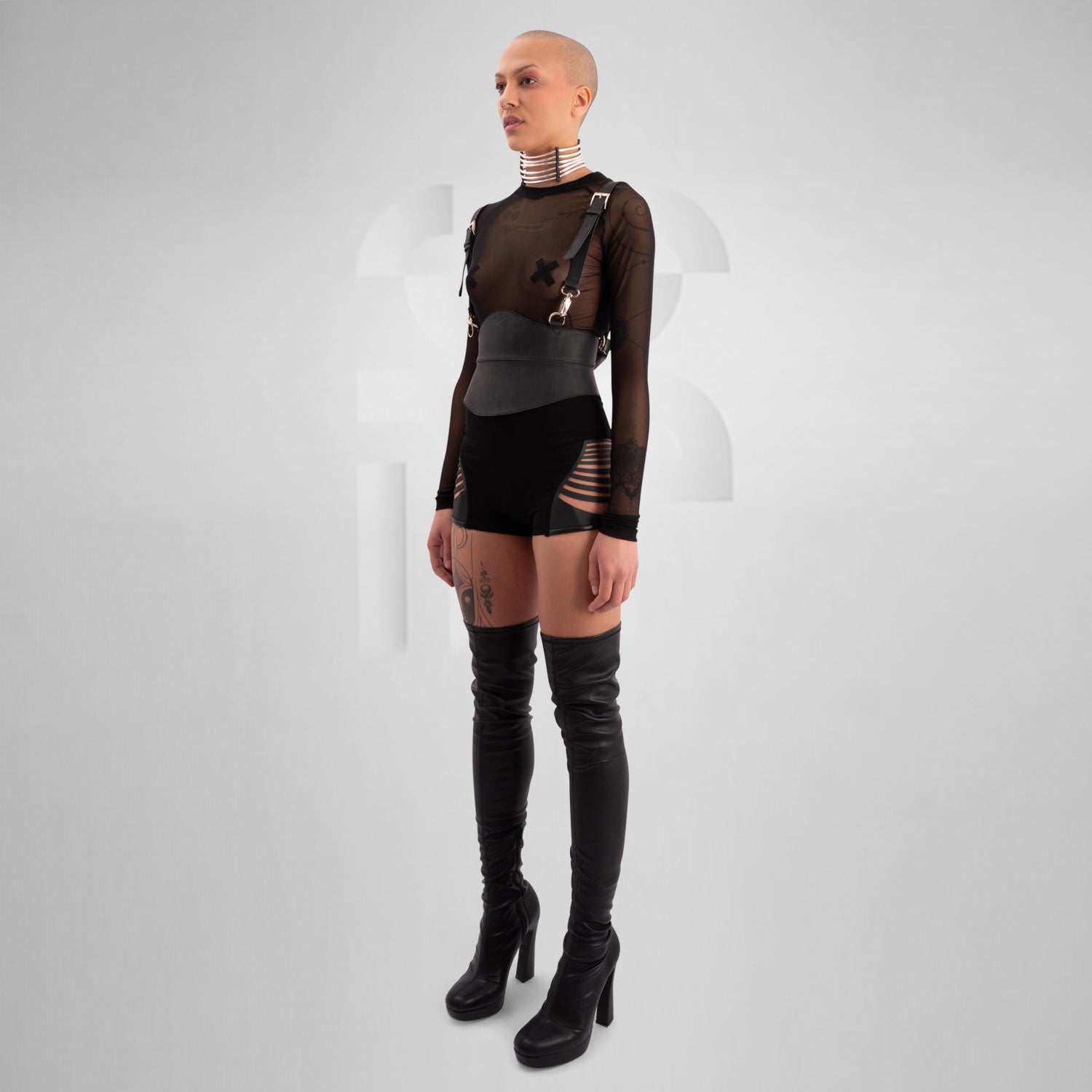 A close-up of a model wearing a sheer black top with decorative straps, featuring a unique leather harness belt and a metallic choker collar, showcasing a bold and edgy fashion statement against a neutral gray background.