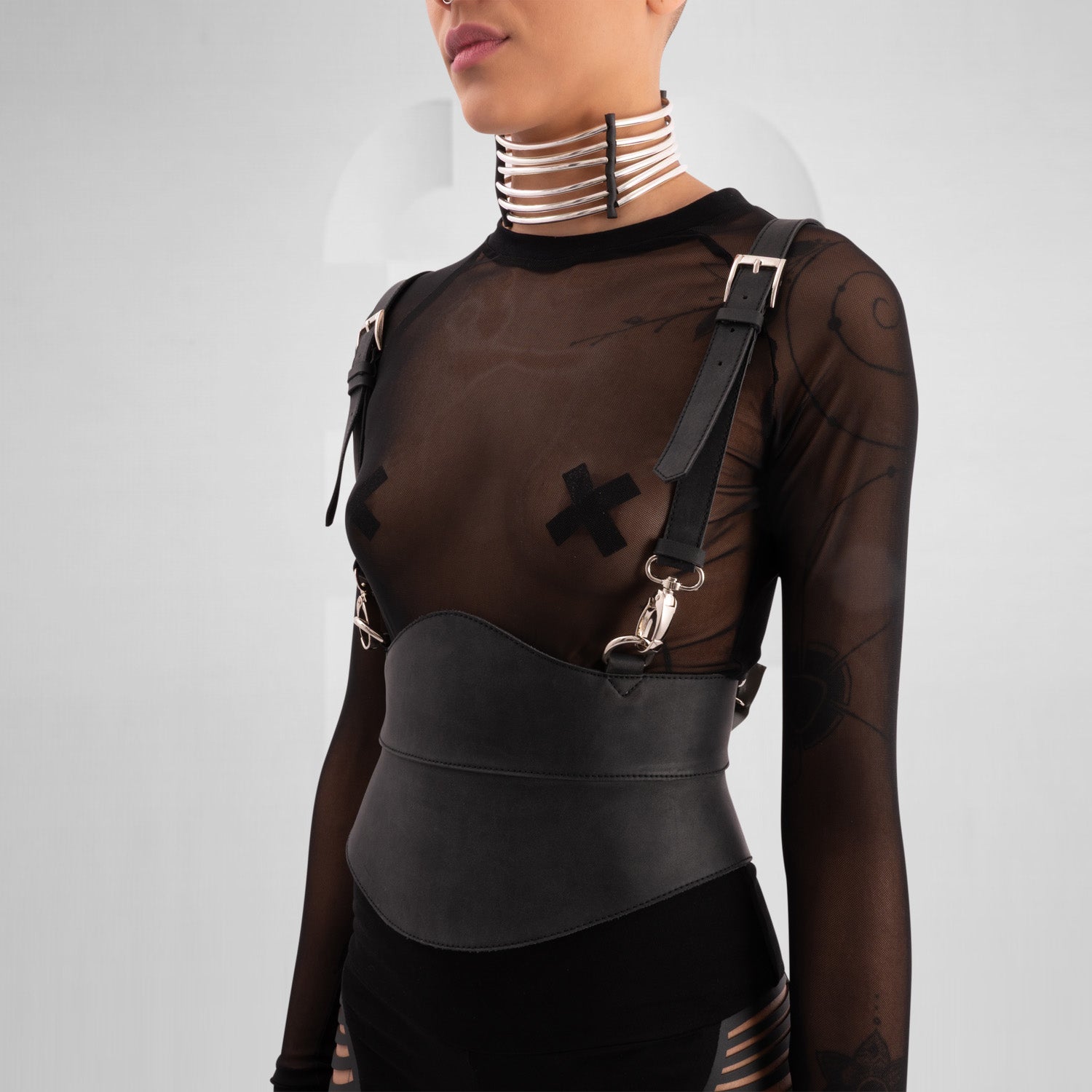 A close-up of a model wearing a sheer black top with decorative straps, featuring a unique leather harness belt and a metallic choker collar, showcasing a bold and edgy fashion statement against a neutral gray background.